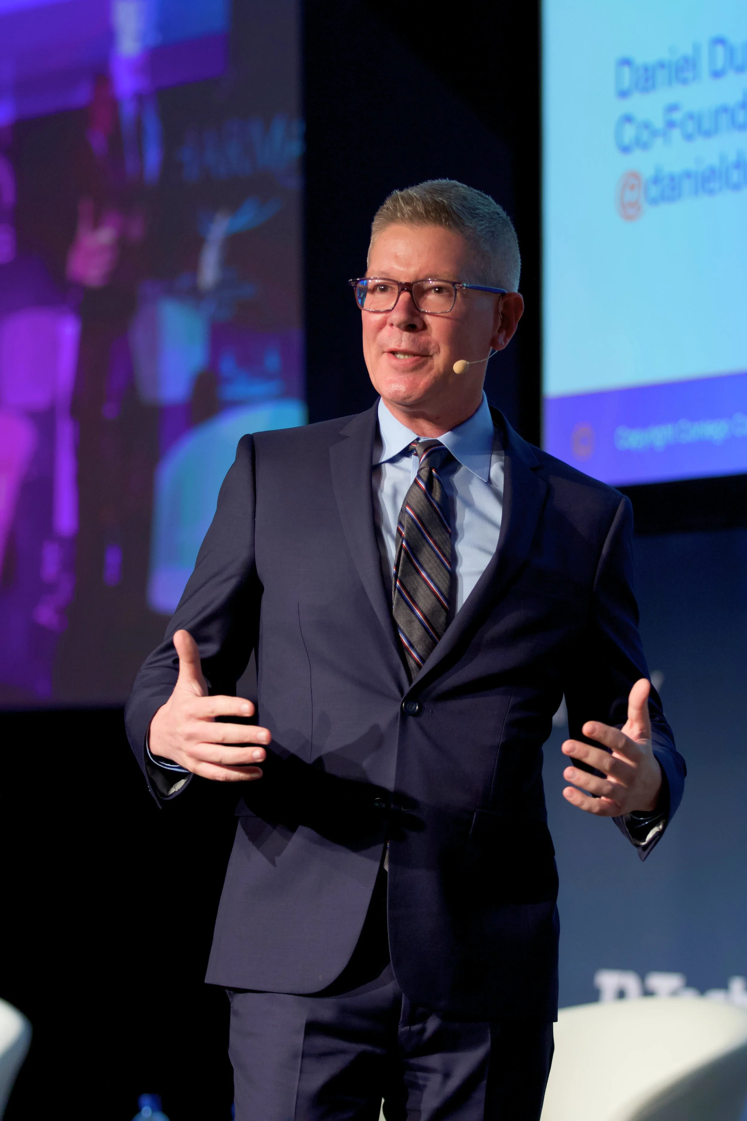 A man in a navy suit, light blue shirt, and striped tie giving a presentation on stage with a blue screen behind him. He is wearing glasses, has short gray hair, and is gesturing with his hands.