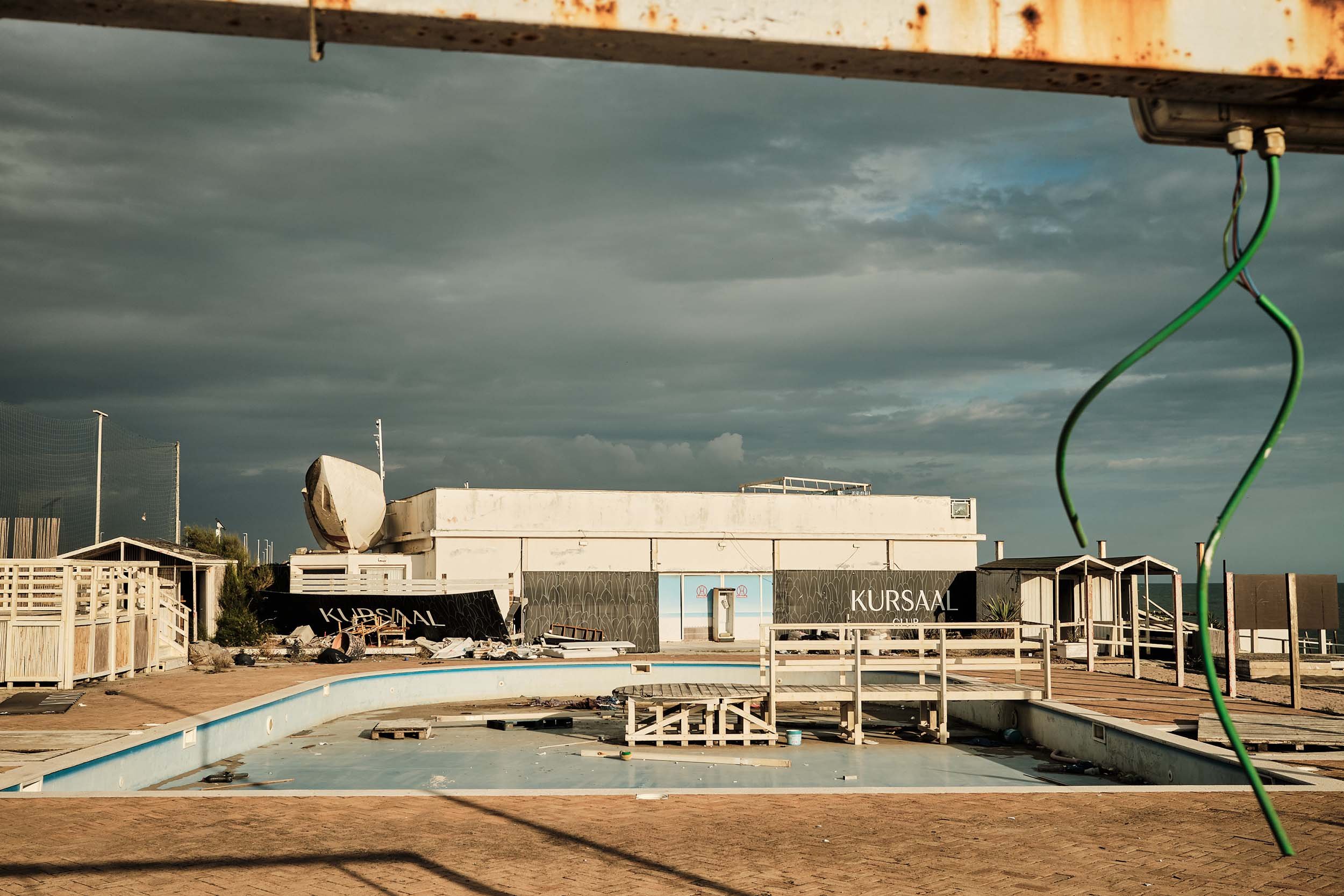 Piscina vuota e macerie al Kursaal di Ostia. In primo piano, sotto un cielo plumbeo, pende un cavo elettrico verde reciso.