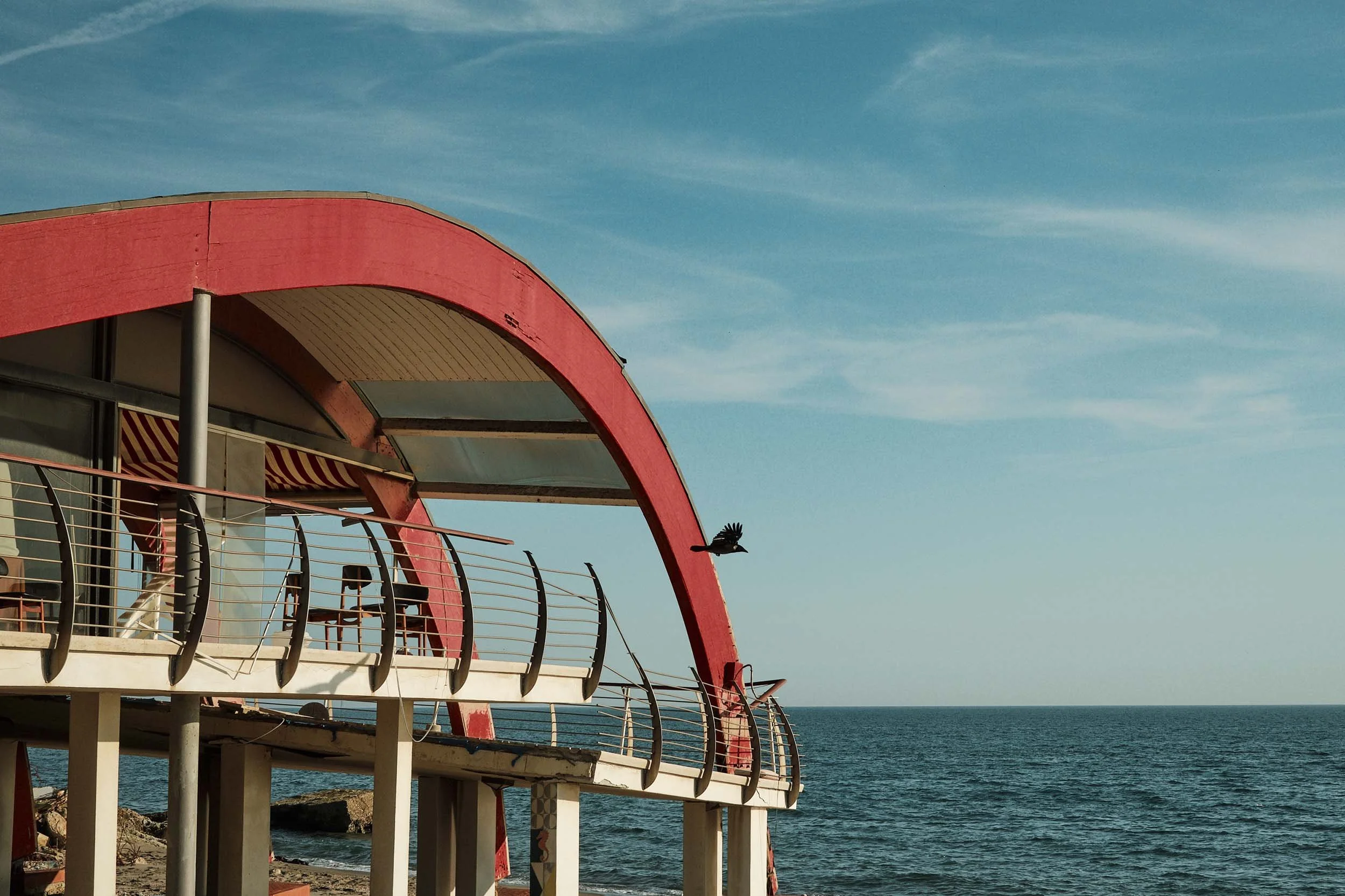 Dettaglio architettonico di uno stabilimento balneare di Ostia, lo Sporting Beach.  La struttura, completamente deserta presenta evidenti segni di abbandono e degrado. la terrazza a sbalzo è staccata di netto. Un uccello nero attraversa il fotogramma