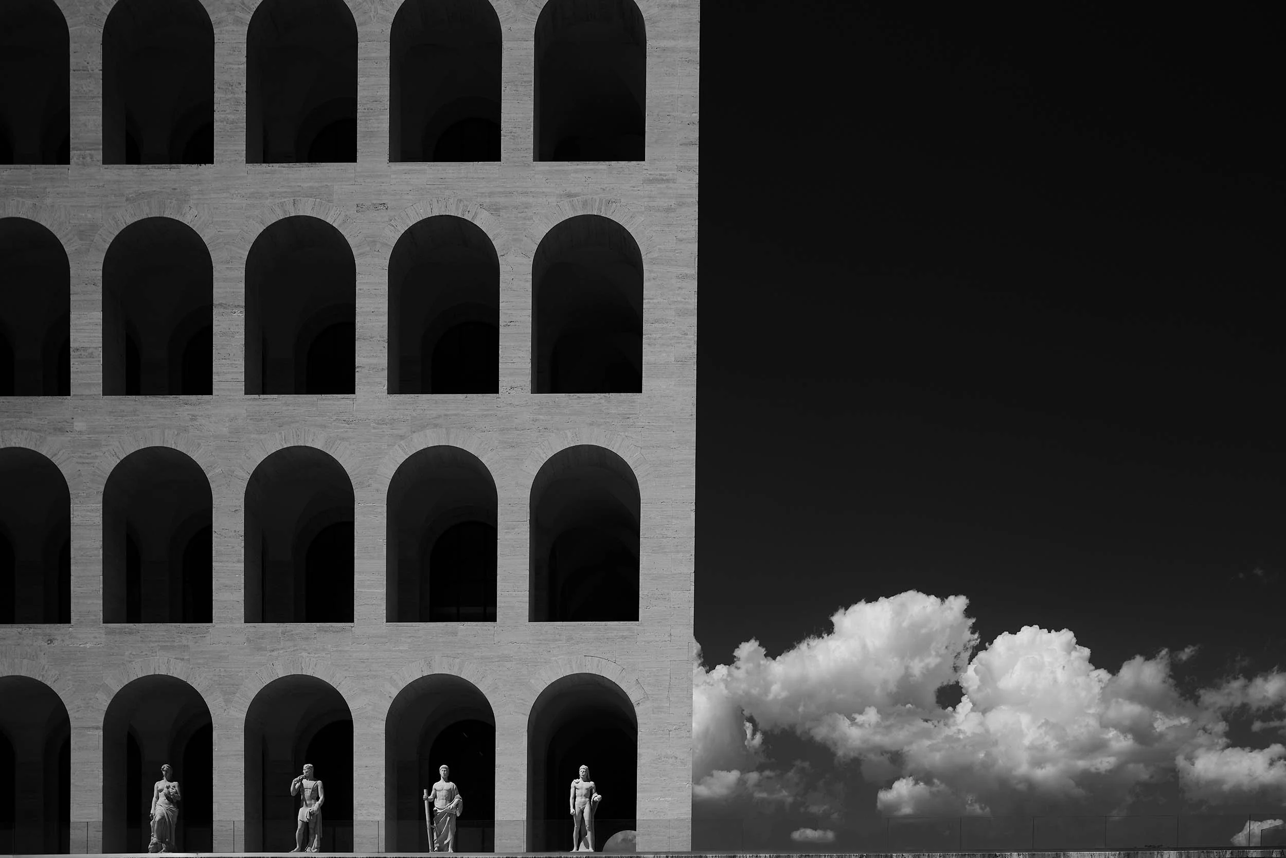 L'architettura del vuoto a Roma: il Palazzo della Civiltà Italiana tagliato a metà da uno spazio negativo assoluto. Le geometrie del travertino e le statue si stagliano contro un cielo nero denso di tensione, spezzato da una nuvola bianca.
