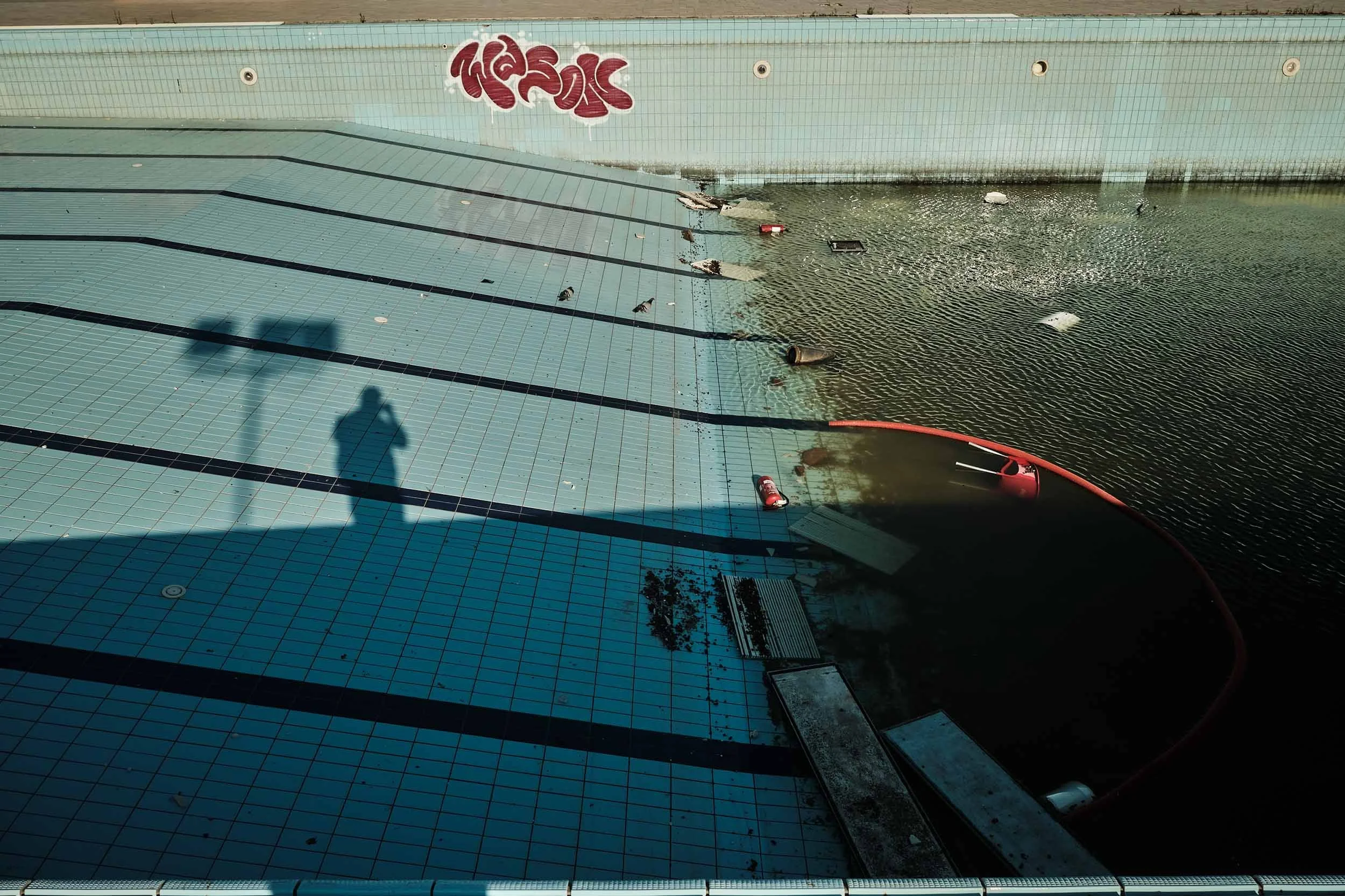 Veduta dall'alto della piscina storica del Kursaal, Ostia. Sulle piastrelle si staglia l'ombra lunga del fotografo.  Sullo sfondo un murale e sulla destra l'acqua stagnante, verdastra e piena di detriti segnalano in modo evidente l'abbandono.