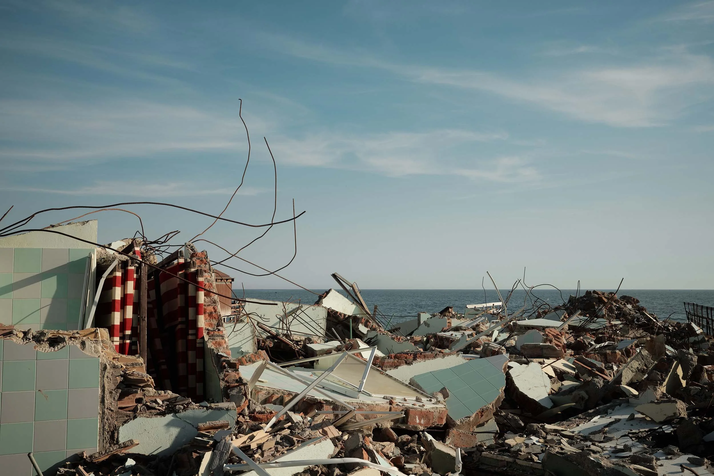 Un cumulo di macerie dello storico stabilimento Sporting Beach sul litorale di Ostia. Cemento armato spezzato, piastrelle rotte e ferri contorti sti stagliano sulla linea del mare, calmo e placido.  Sulla sinistra, i resti di alcune sdraie a righe.