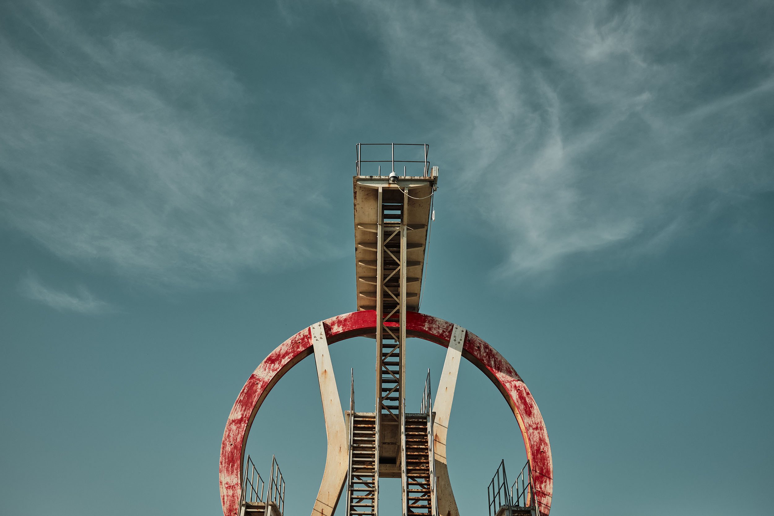 Inquadratura simmetrica, dal basso, dello storico trampolino del Kursaal di Ostia, capolavoro di Pierluigi Nervi.  La monumentale struttura bianca e rossa, stagliata contro il cielo, mostra segni di abbandono, ruggine, sfaldamento e incuria.