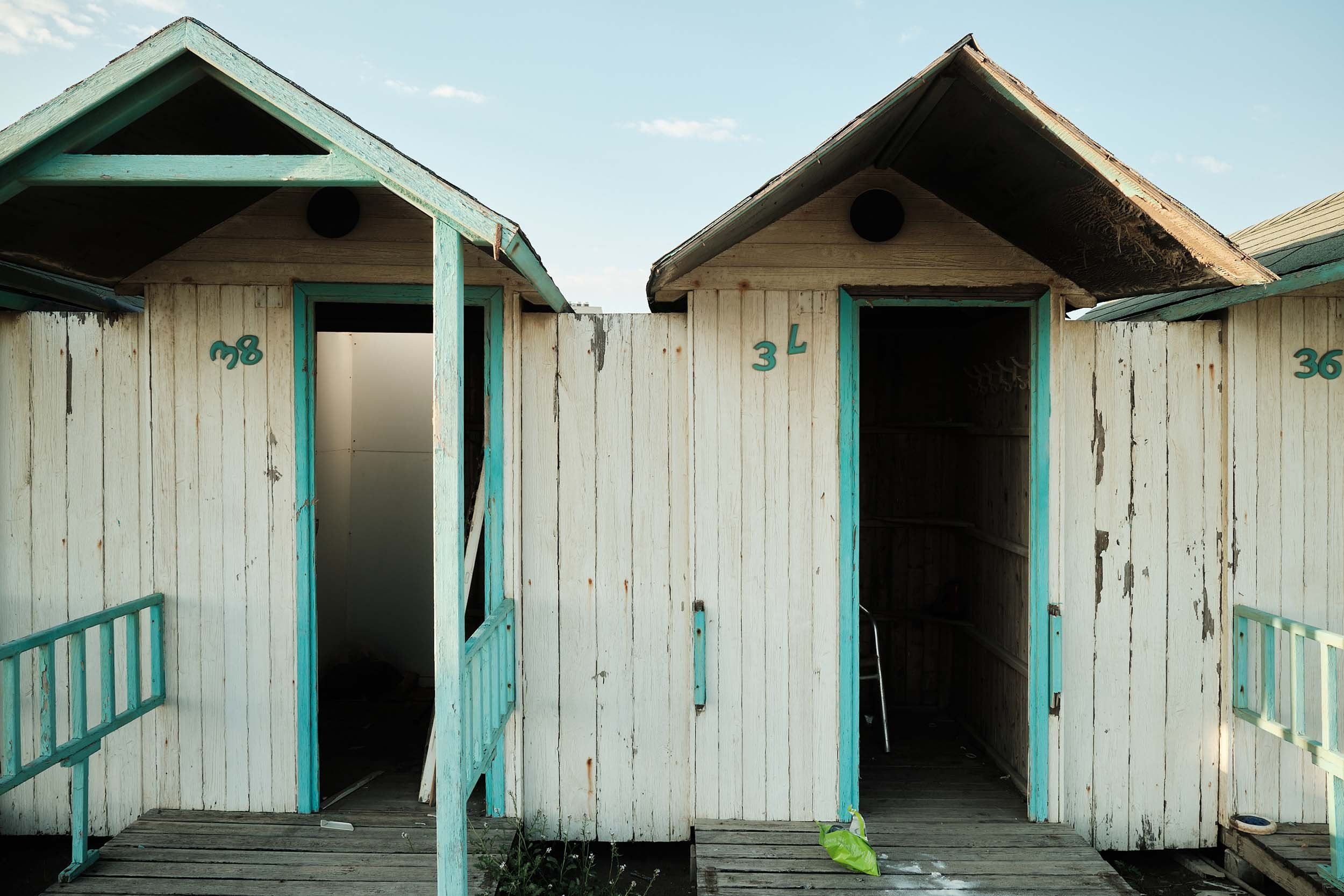 Vecchie cabine balneari in legno bianco e azzurro a Ostia. Vernice scrostata, ingressi aperti e interni inghiottiti dal buio.