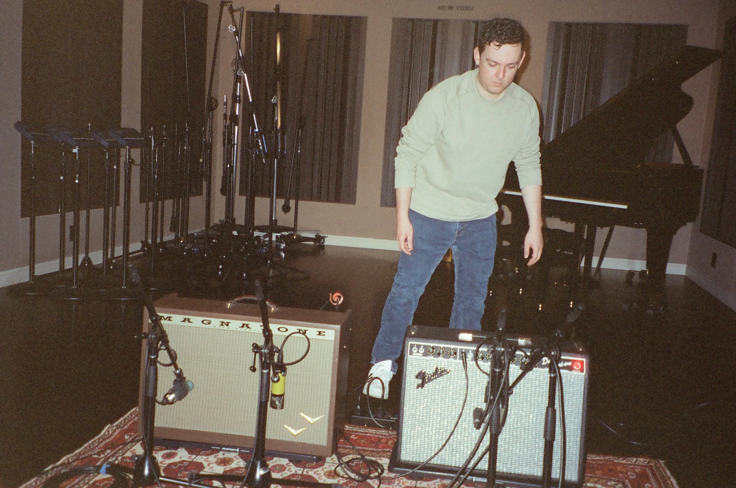 Two guitar amps with microphones in a recording studio with a sound engineer standing behind mid-motion, a piano and microphone stands staged behind