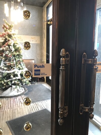 Picture of double doors that have vertical handles with a sticker sign nearby that says “push.”