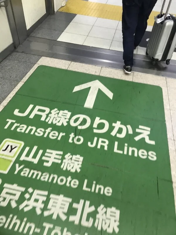 Picture of a sign on the floor at a train stop directing people to a specific line and transfer area in multiple languages.