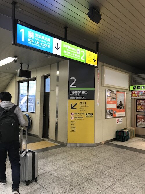 Picture of a sign on a wall and hanging from the ceiling directing people to different train lines at a train stop, both in multiple languages.