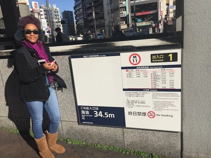 Picture of me standing next to a train stop sign showing the train departure information in Japanese and English.