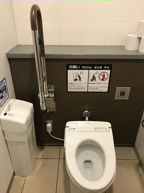 Picture of a clean stall containing a toilet, trash bin, and an assisting metal bar.