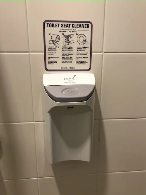 Picture of a dispenser with a sign titled “Toilet Seat Cleaner” above it, containing 3 main directions on how to use it in multiple languages.