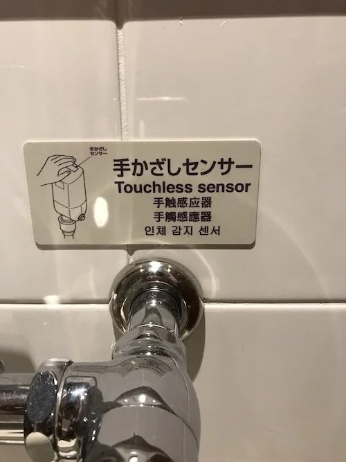 Picture of a sign above a toilet that reads “Touchless sensor” in multiple languages, along with an illustration of a hand hovering over a device.