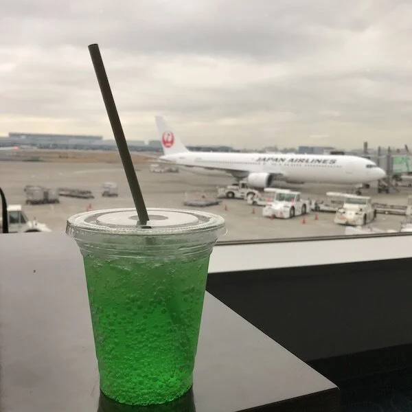 Picture of a cup of the green Melon Fanta on a table with a parked Japan Airlines plane in the background.