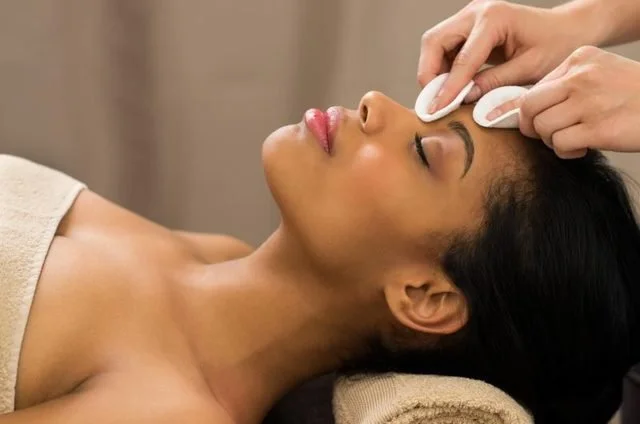 A woman receiving a facial treatment, with a beauty professional applying a product to her forehead using a cotton pad.