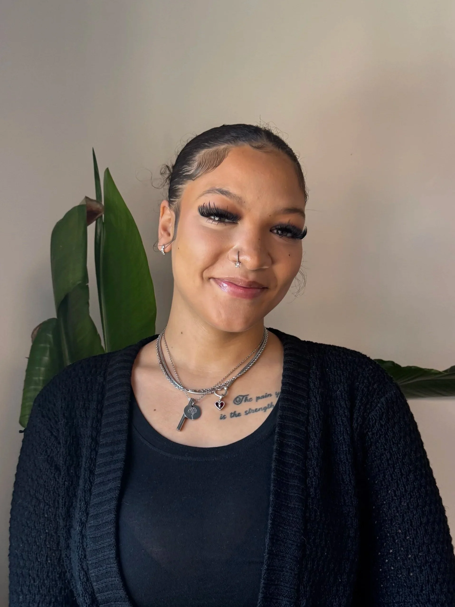Young woman with dark hair slicked back, wearing jewelry including layered necklaces, earrings, and a septum piercing, smiling in front of a beige wall with green plant leaves in the background.