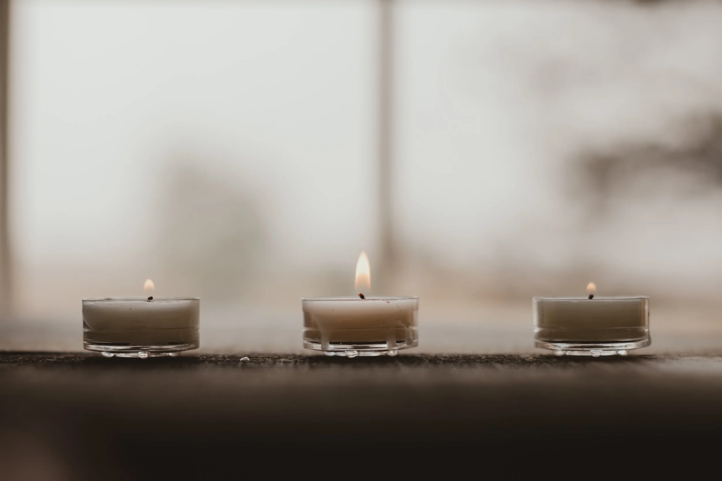 Three small lit tea light candles placed on a dark surface with a blurred background.