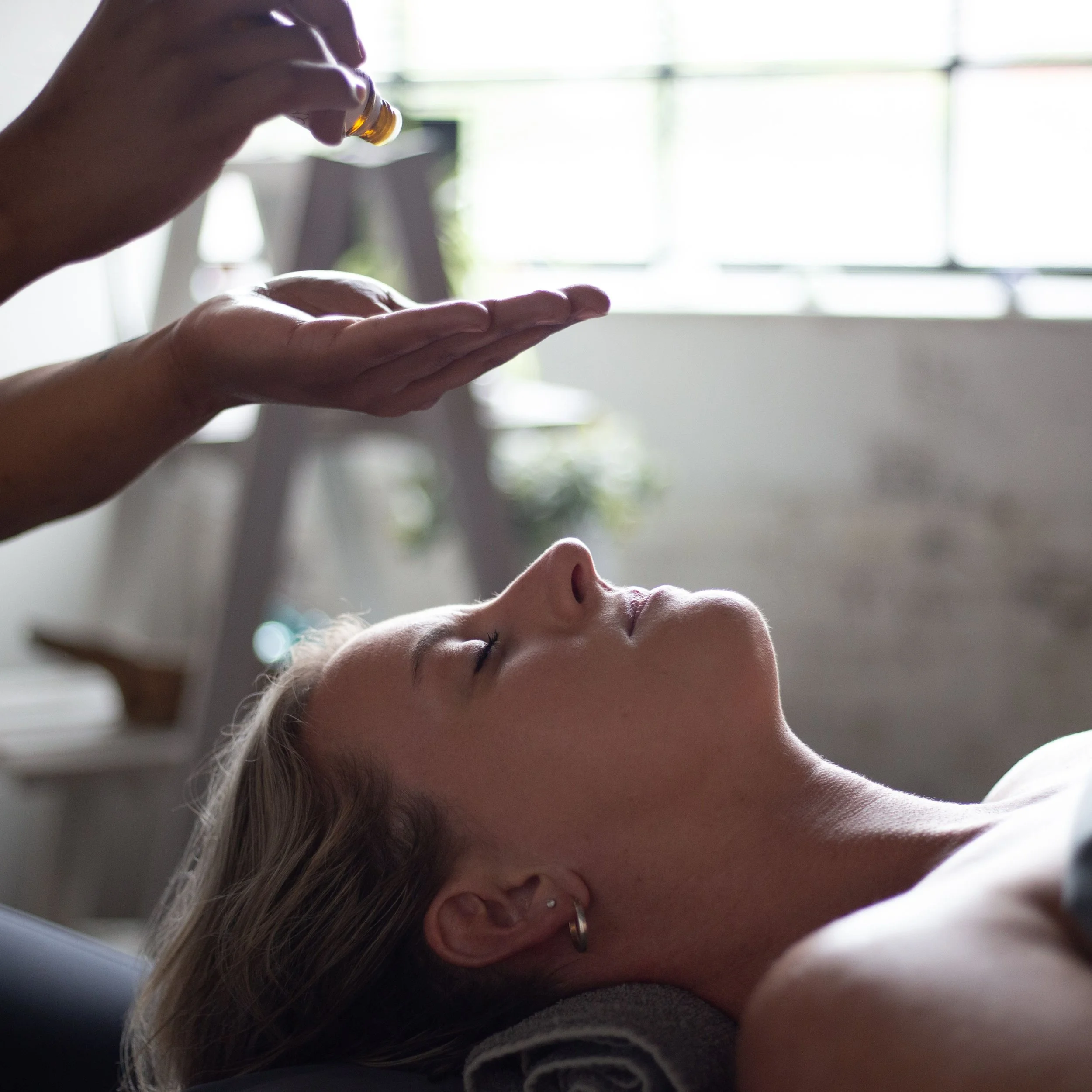 A woman receives an essential oil massage or treatment while lying down indoors near a window.