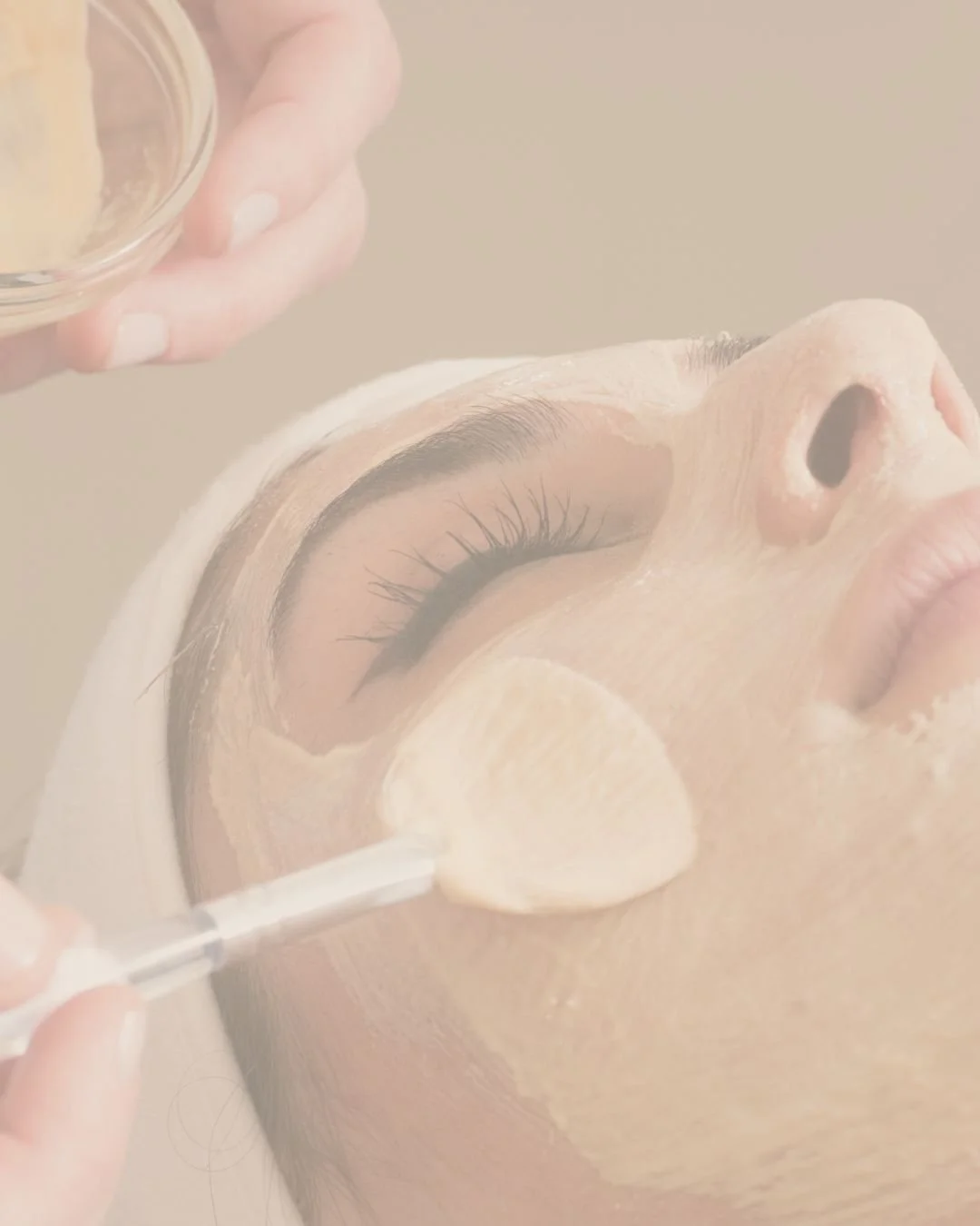 A person receiving a facial treatment with a facial mask applied, and a beauty professional applying a product with a brush.