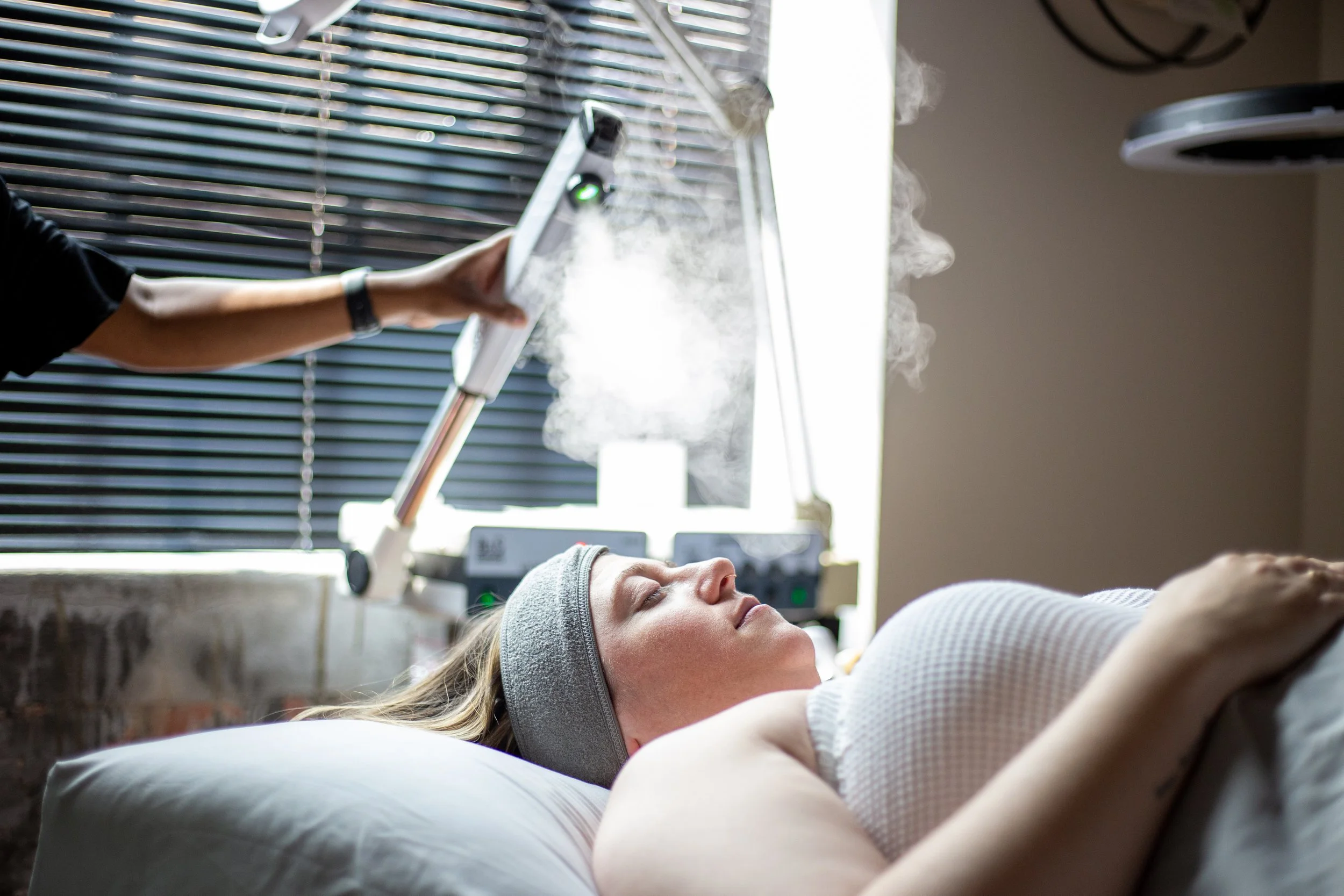 Woman lying down with eyes closed, receiving a facial treatment from a technician using a steam device, in a spa or skincare clinic.