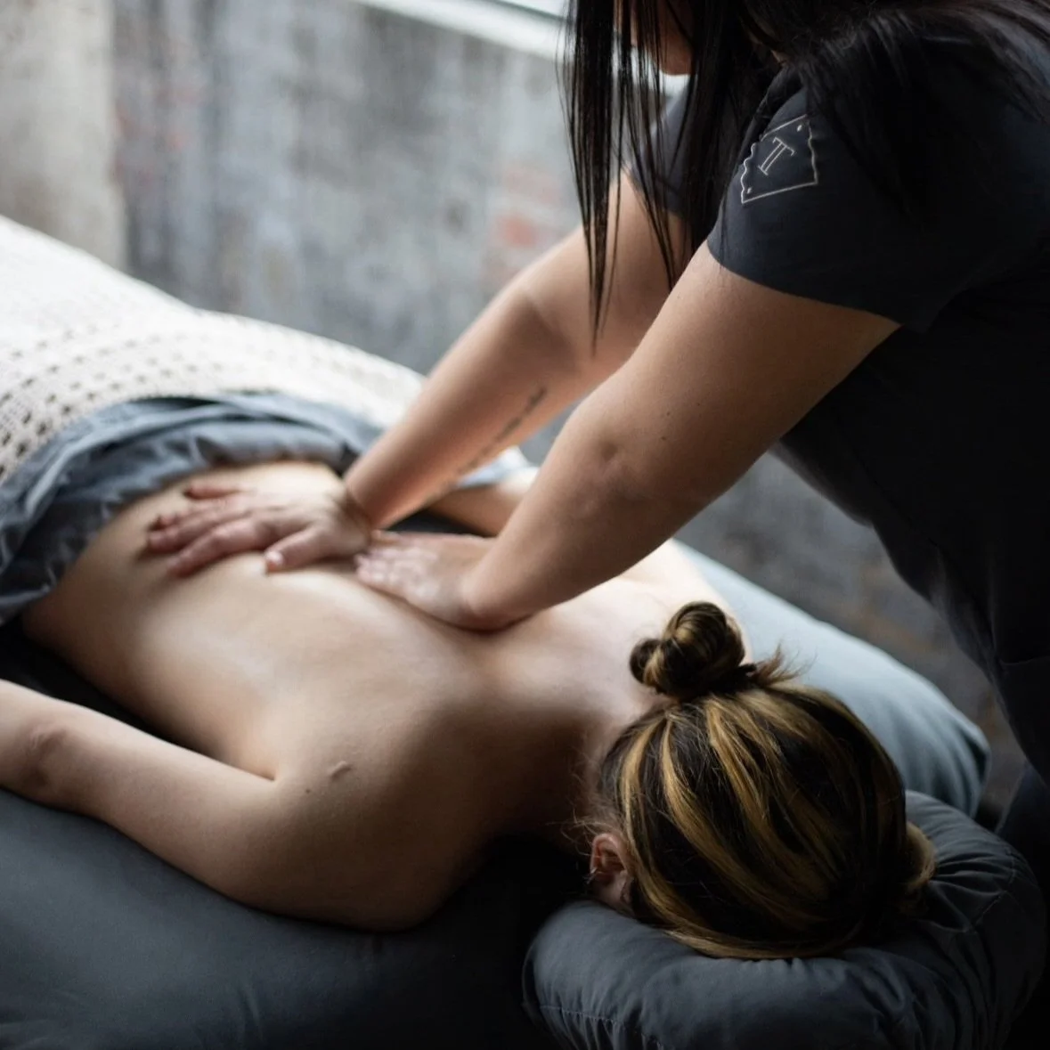 A woman gives a massage to a person lying face down on a massage table in a dimly lit room.