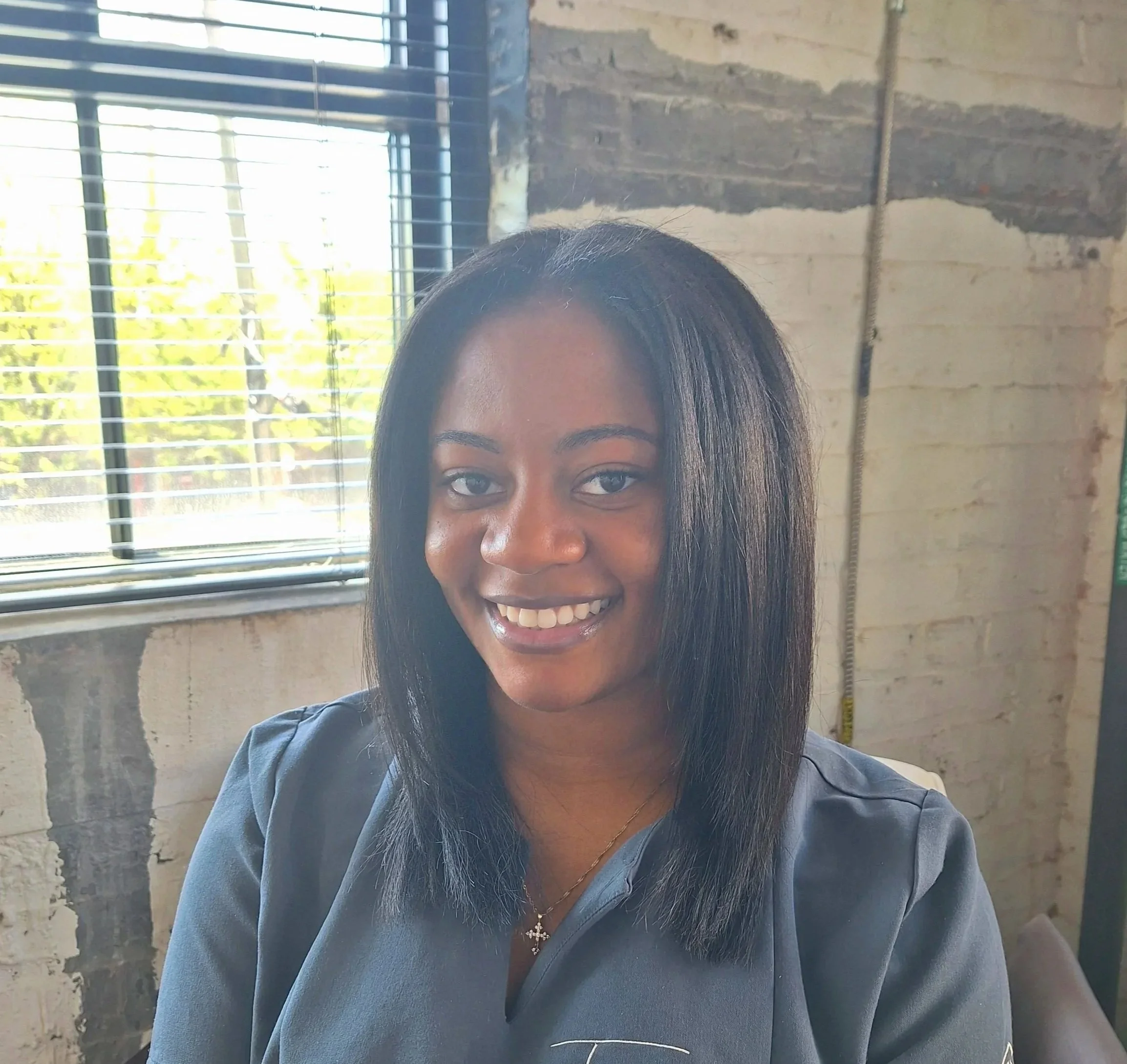 A woman smiling at the camera, wearing a dark gray shirt with the words "Tranquil Traditions" embroidered on it, sitting indoors with a window and white brick wall in the background.