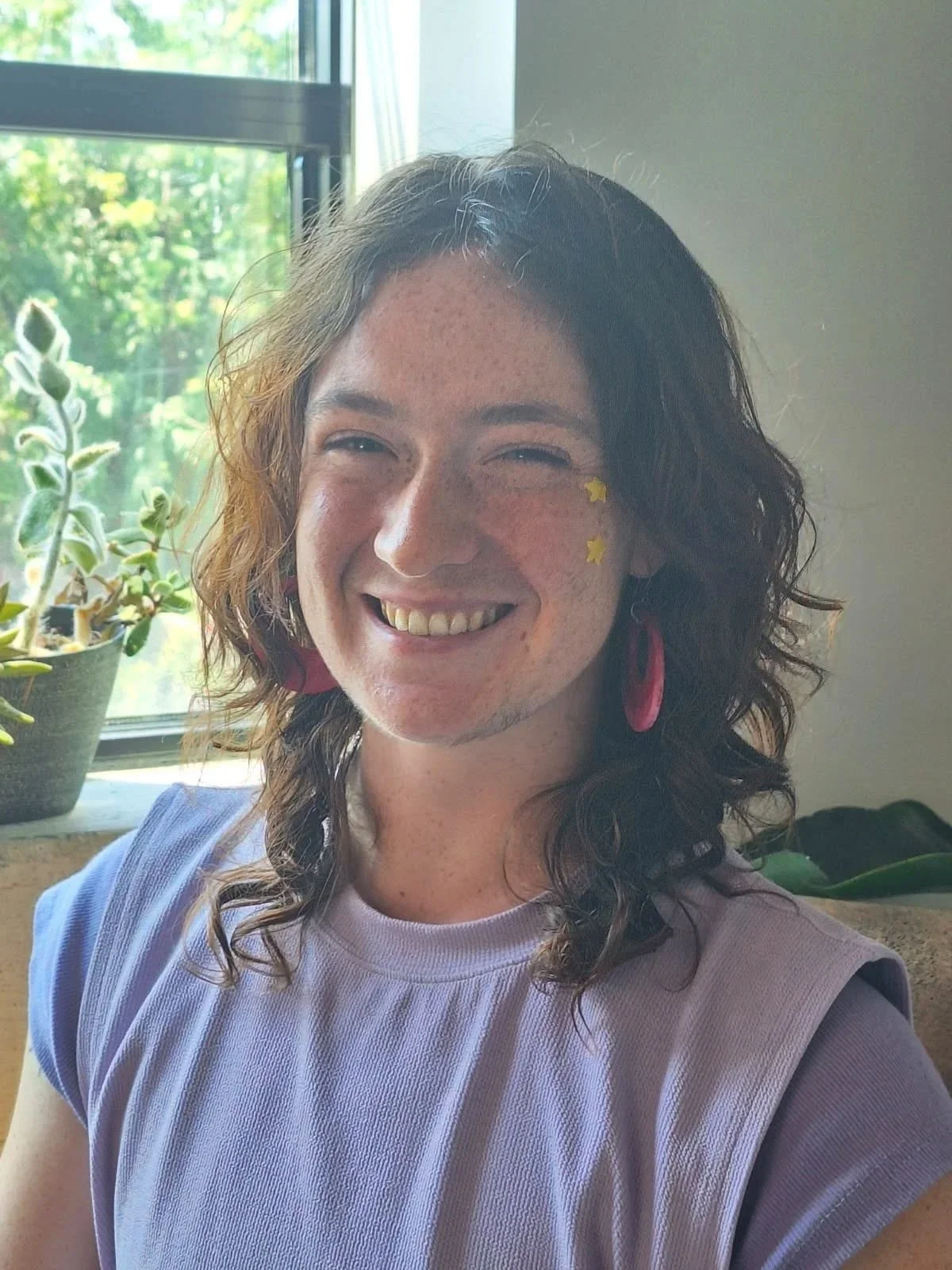 Smiling woman with curly hair, wearing pink earrings, face stickers, in front of window with plants.