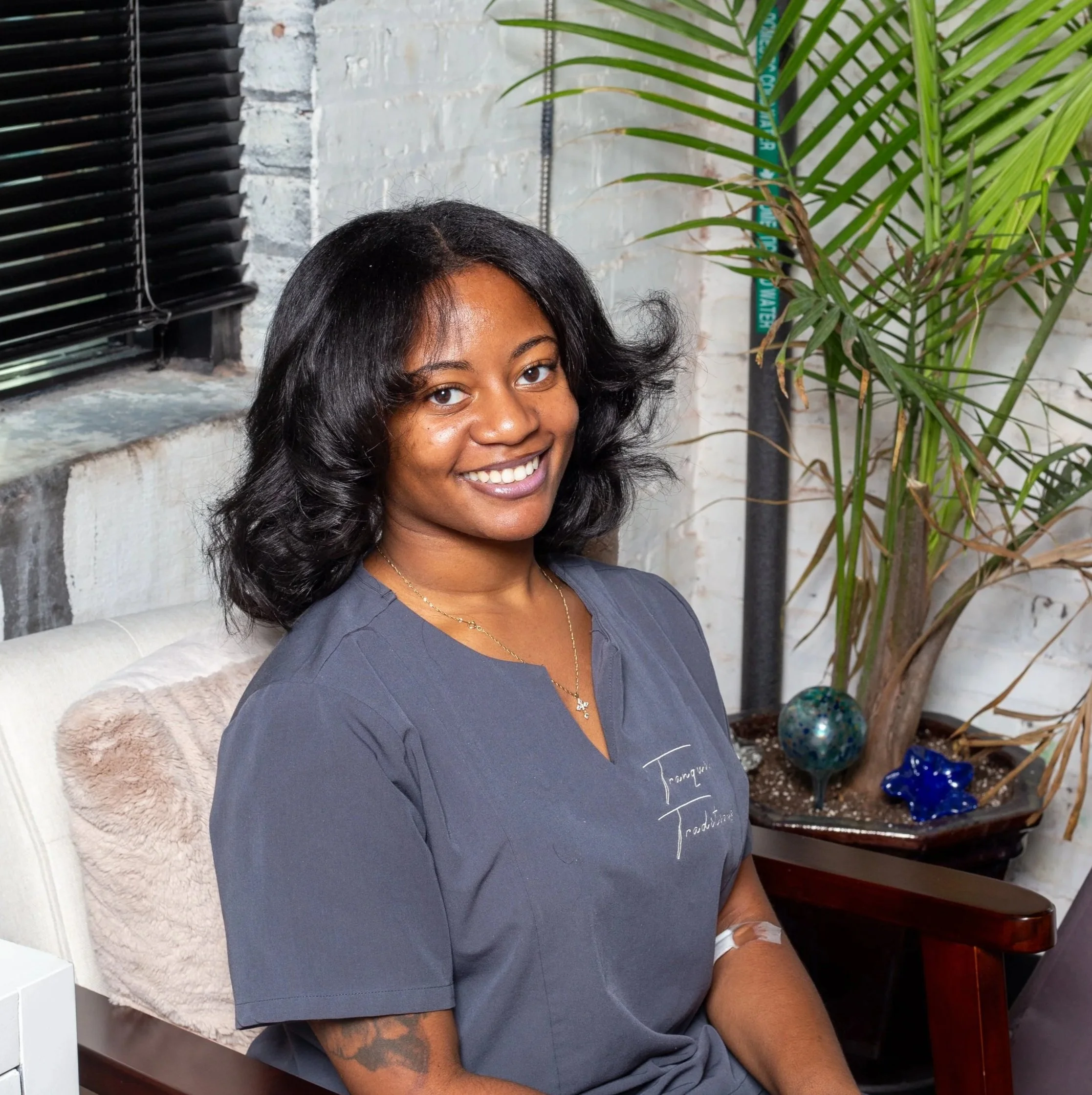 A woman smiling at the camera, wearing a dark gray shirt with the words "Tranquil Traditions" embroidered on it, sitting indoors with a window and white brick wall in the background.