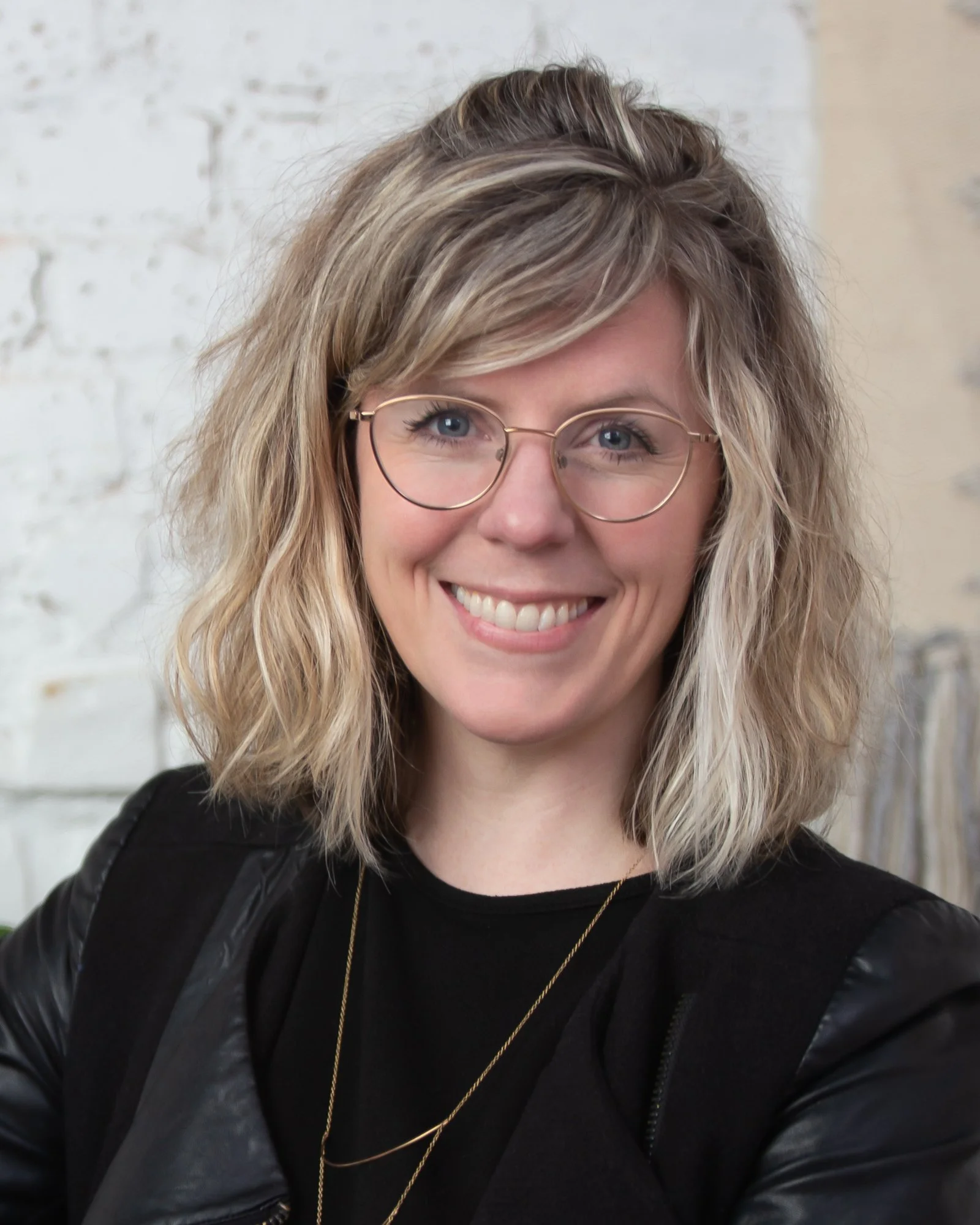 A woman with shoulder-length wavy blonde hair, wearing glasses and a black jacket, smiling at the camera.