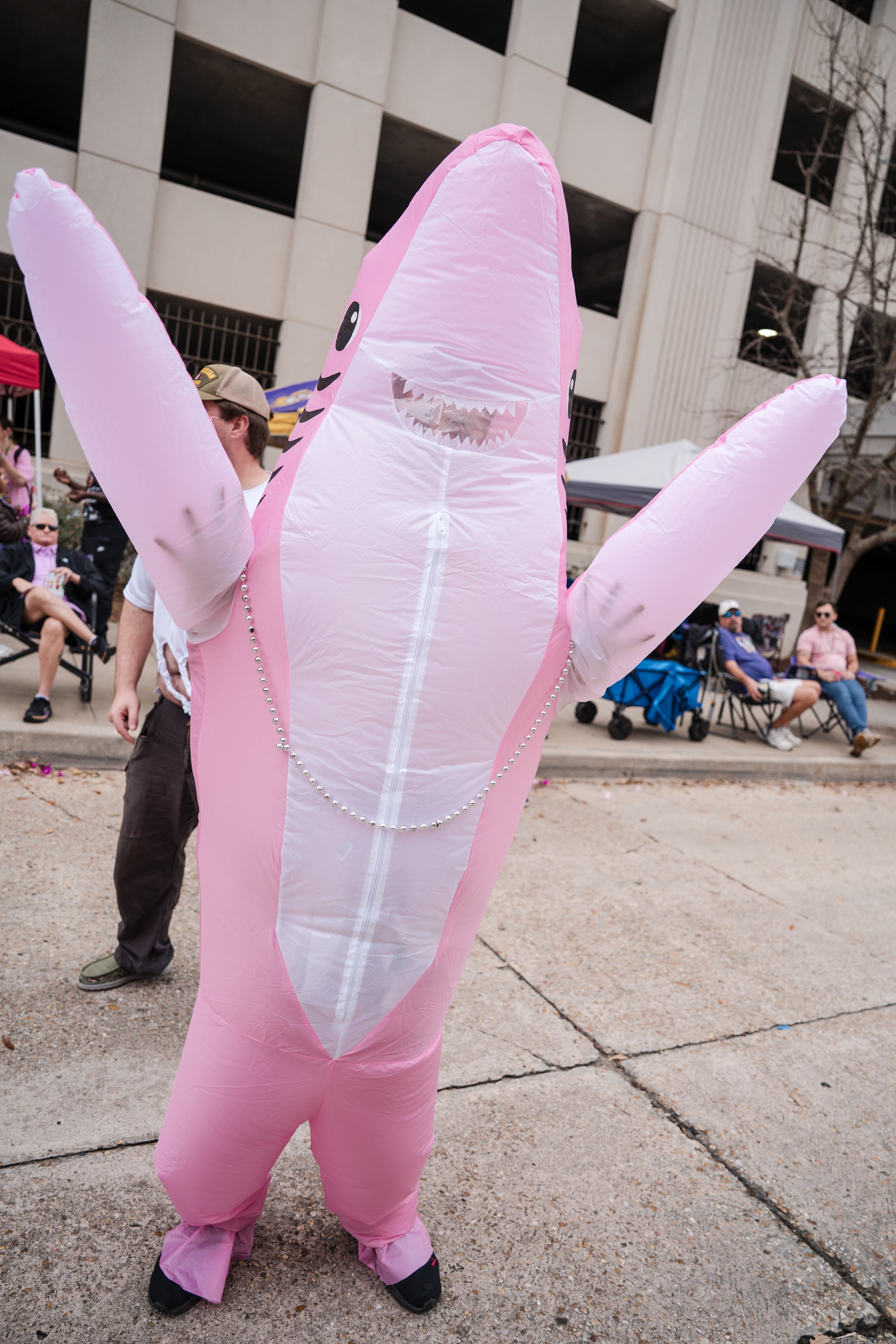 Person dressed in a pink inflatable shark costume with arms raised at an outdoor event with seated spectators in the background.