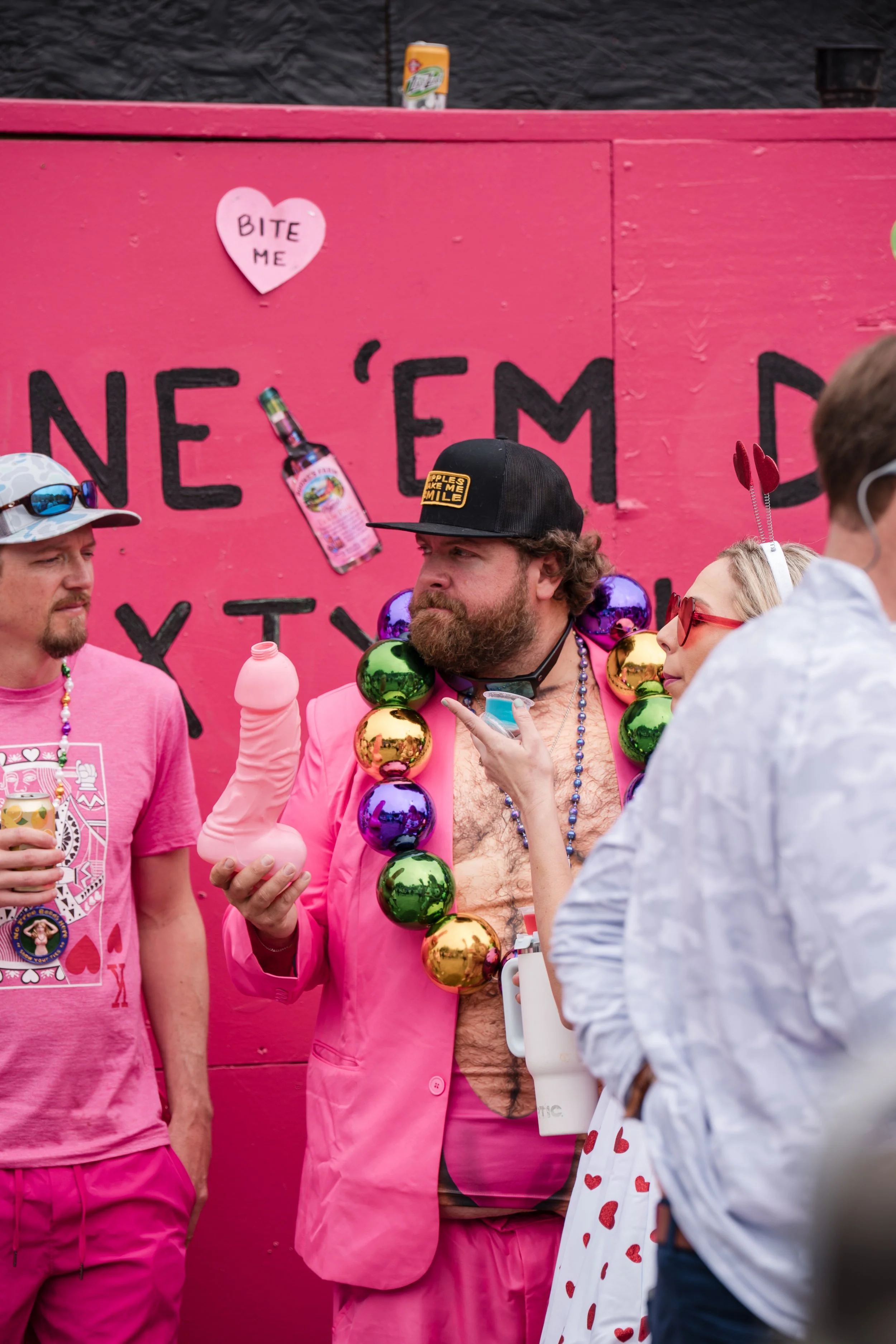 People dressed in pink with colorful beads and accessories, celebrating at an event with a pink backdrop that reads "N'Em D" in the background. The man in the center holds a pink inflatable sex toy and has a necklace of large colorful ornaments aroun