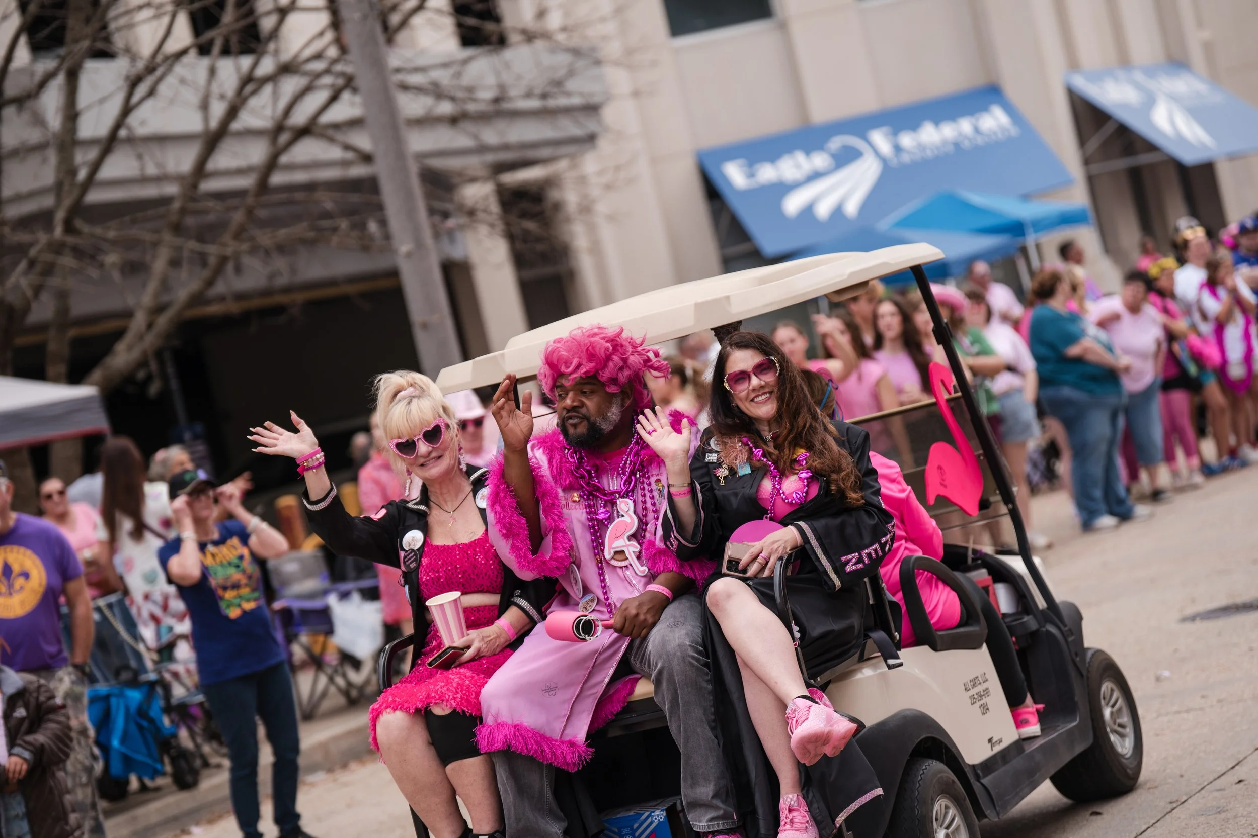 Three people riding in a golf cart dressed in pink and black, surrounded by a crowd at a parade. One woman with blonde hair and pink sunglasses sits holding a striped cup. The man in the middle has pink curly hair and a pink feathered accessory. The 