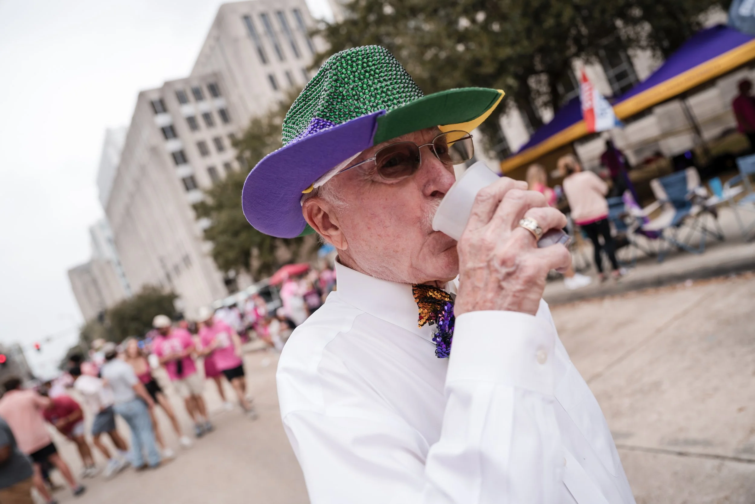 Elderly man with sunglasses and a colorful beaded hat drinking from a paper cup at an outdoor event, with people and tents in the background.