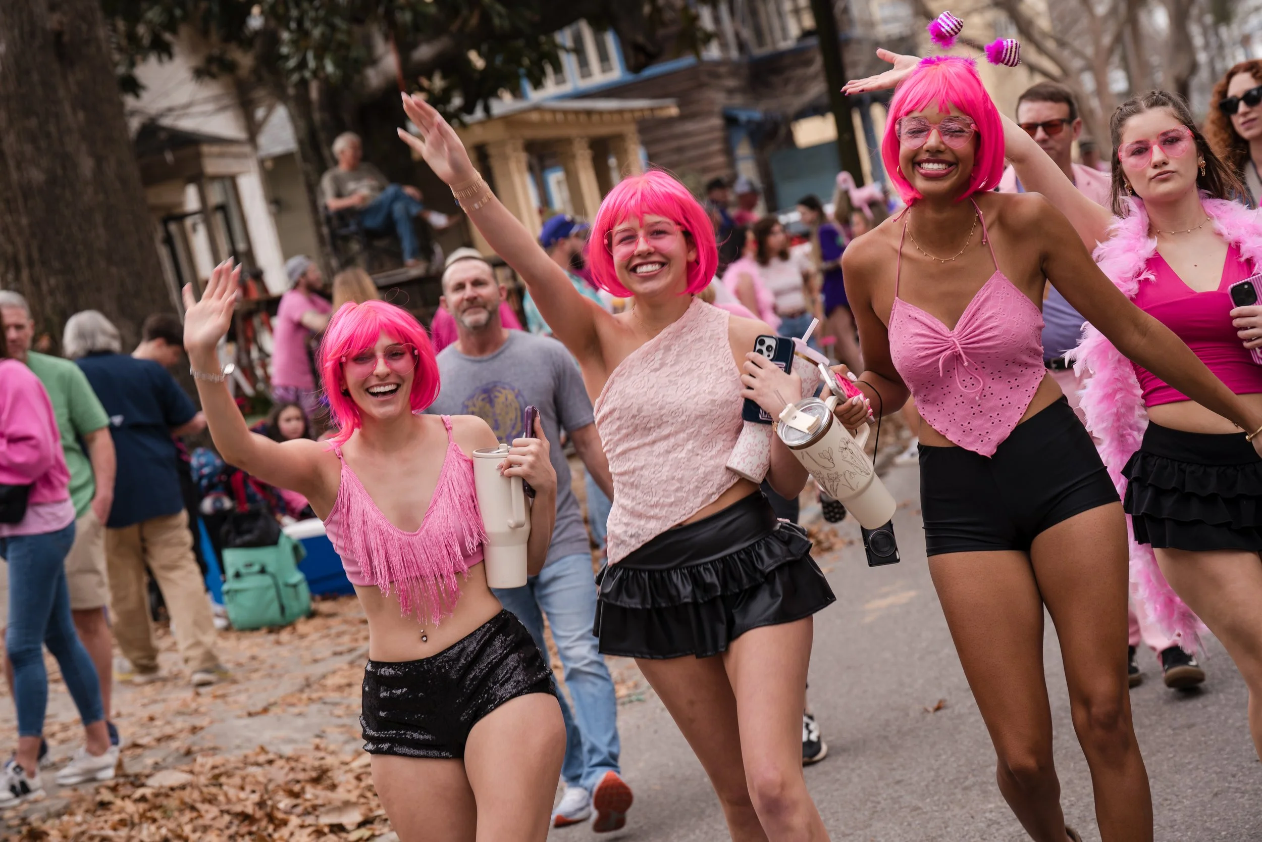 Three women in pink wigs and outfits smiling and waving during a parade or festival with other people and houses in the background.