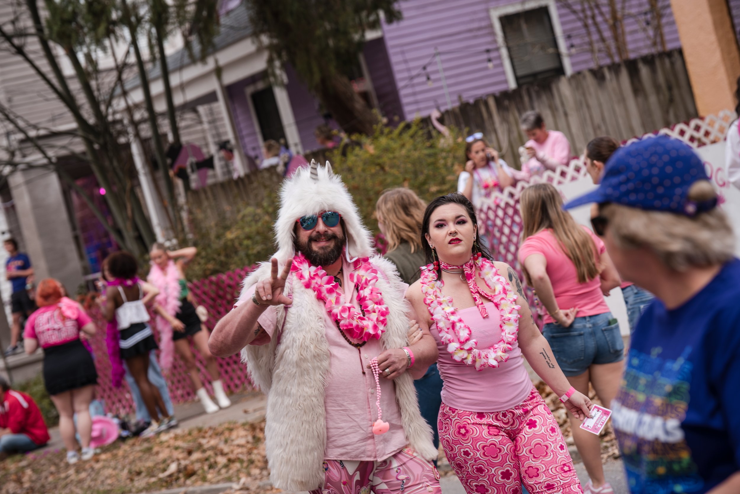 People dressed in pink and wearing leis at a daytime outdoor event, with some in costume including one person in a white furry unicorn hat and glasses, and others in casual and themed attire, in a neighborhood street with houses and trees in the back