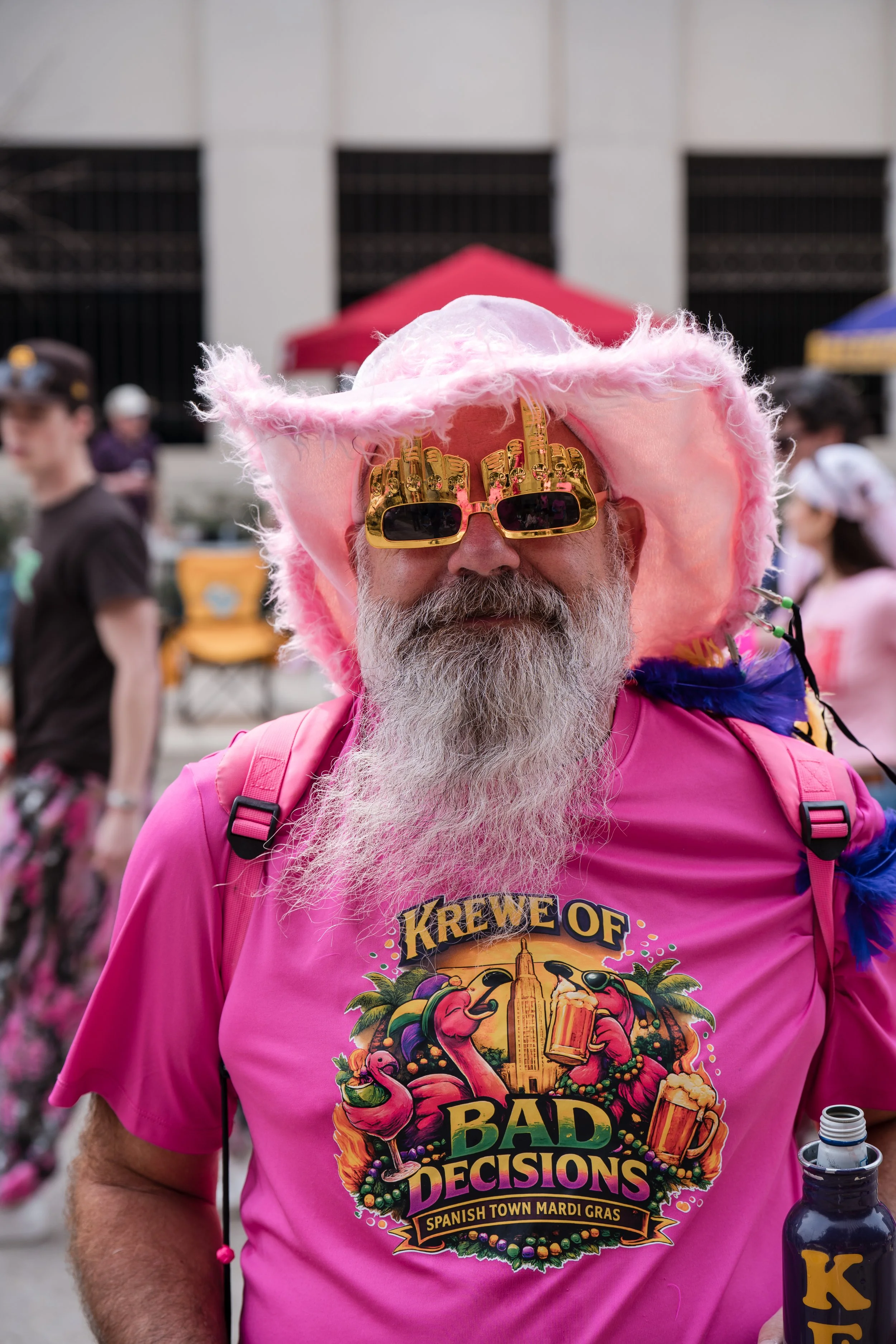 A man with a long gray beard wearing a pink hat, sunglasses, and a bright pink T-shirt with a colorful design. The design features flamingos, drinks, and the text "Krewe of Bad Decisions" and "Spanish Town Mardi Gras." He is outdoors during a festive