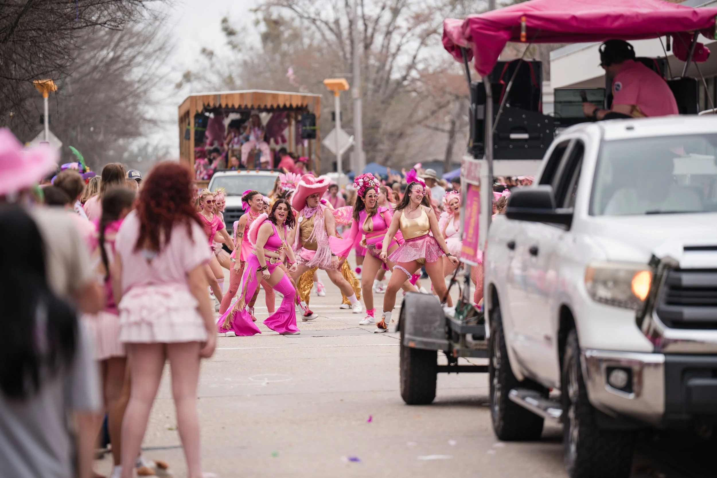 Participants in a parade dressed in pink costumes marching and dancing, with a float in the background and spectators lining the street.
