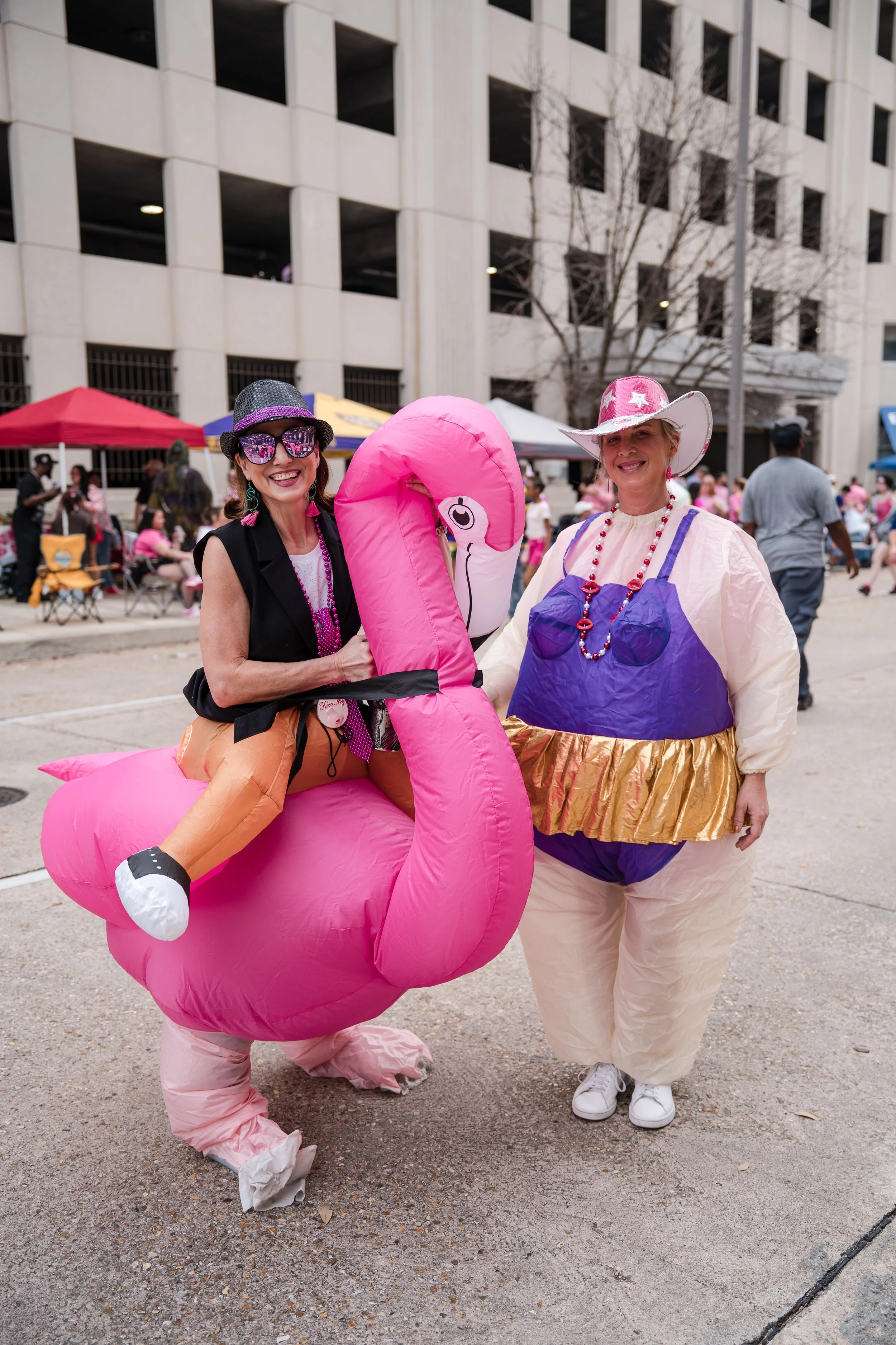 Two women in colorful costumes at a parade, one riding an inflatable pink flamingo, and the other dressed in a whimsical outfit with a large hat, on a city street with onlookers and tents in the background.
