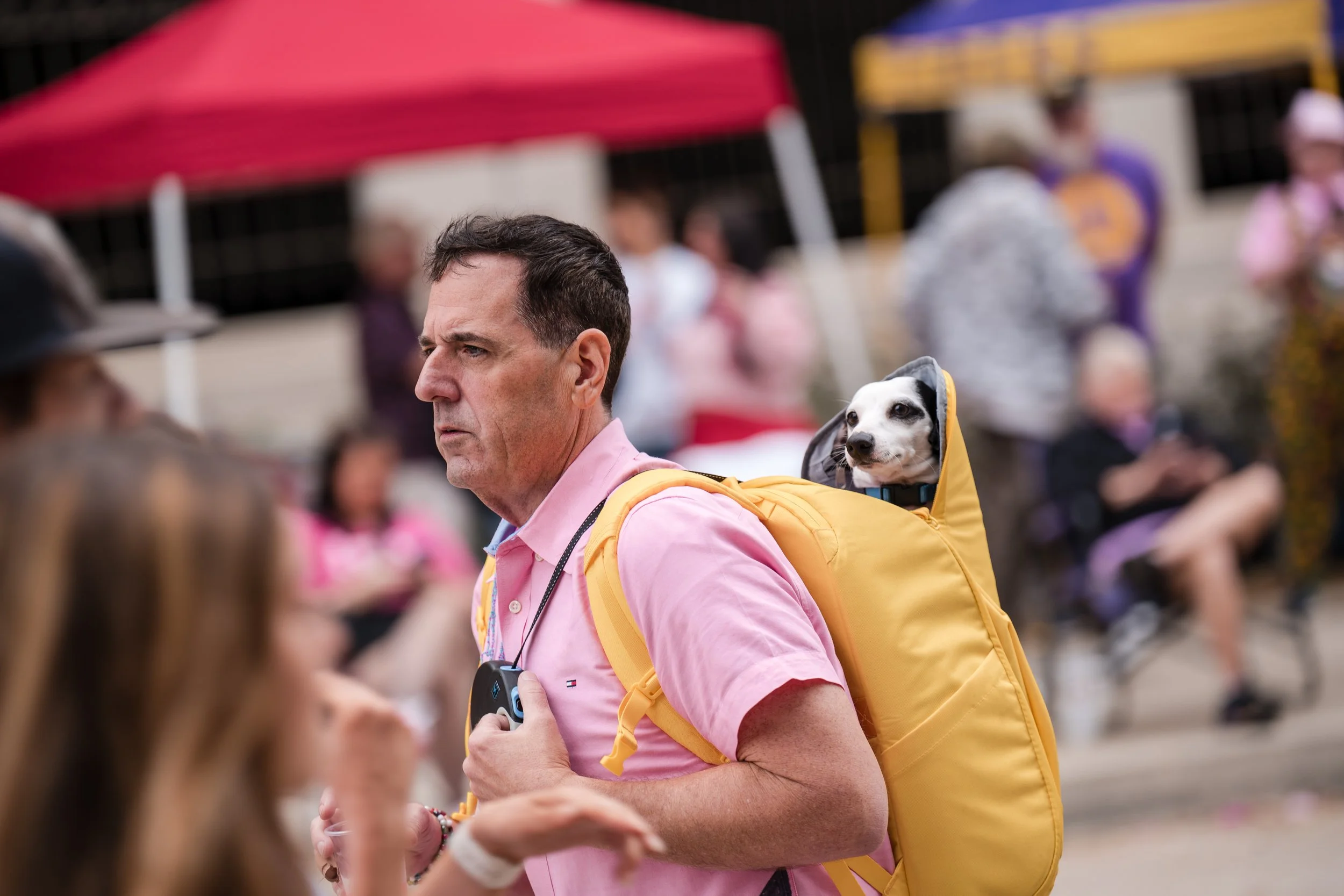 A man wearing a pink shirt carries a yellow backpack with a small dog in a backpack dog carrier. The dog looks out of the carrier, and people are gathered in the background at an outdoor event.