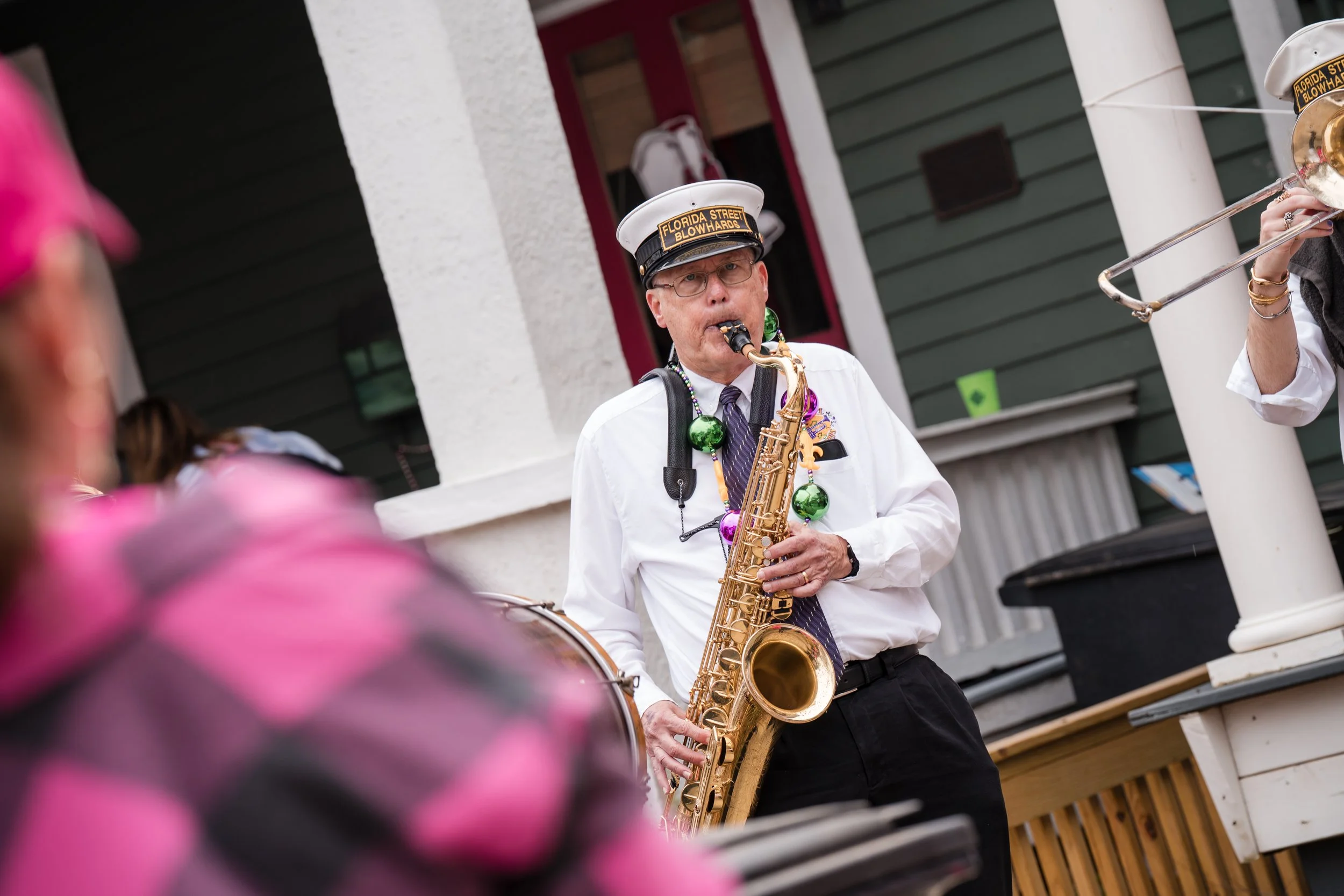 A man playing a saxophone at an outdoor event, wearing a white shirt, purple tie, glasses, and a hat that says 'Florida Street Blowhards,' with colorful Mardi Gras beads around his neck.