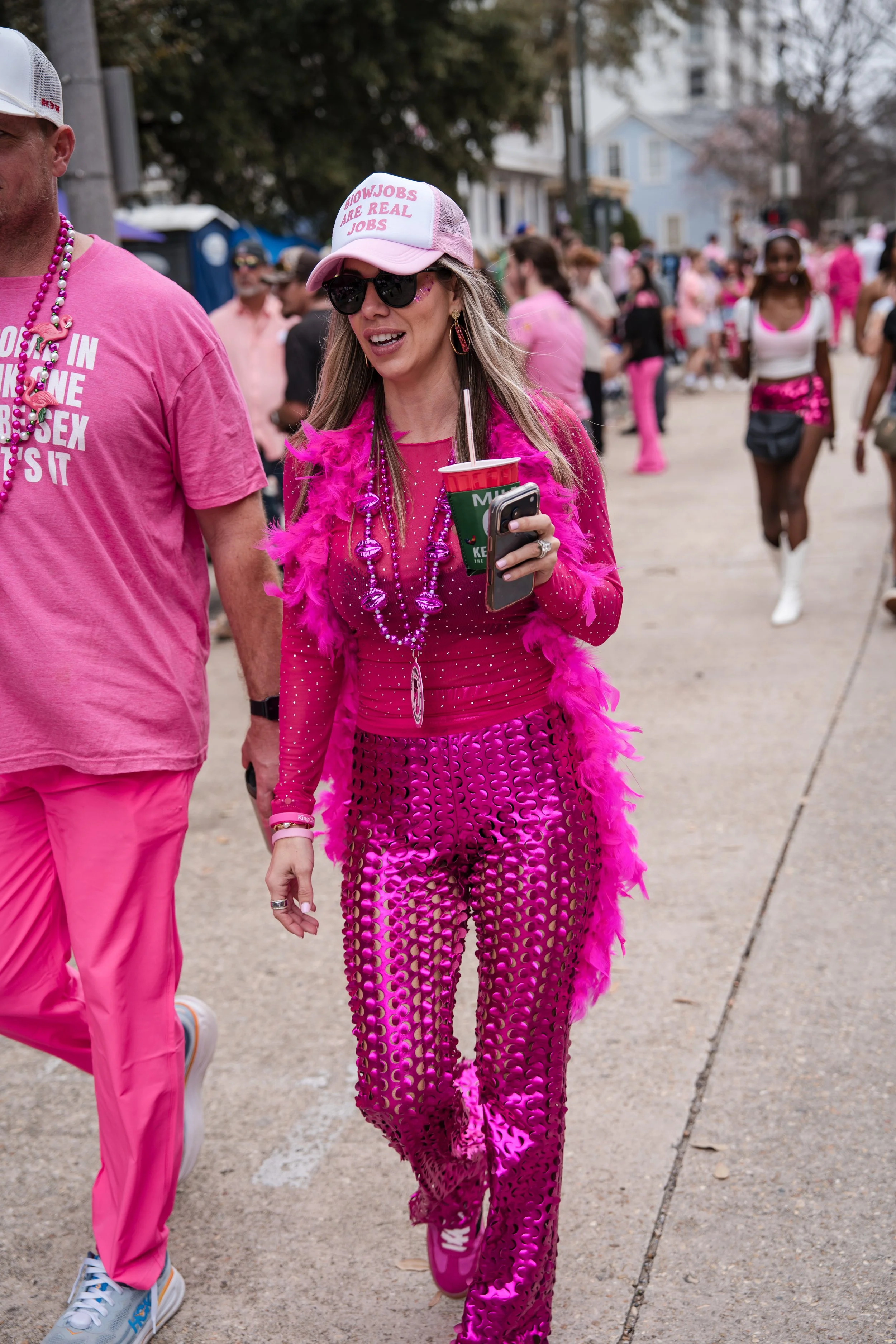 Woman wearing pink, sequined, and feathered clothing, walking outdoors during a parade or festival.