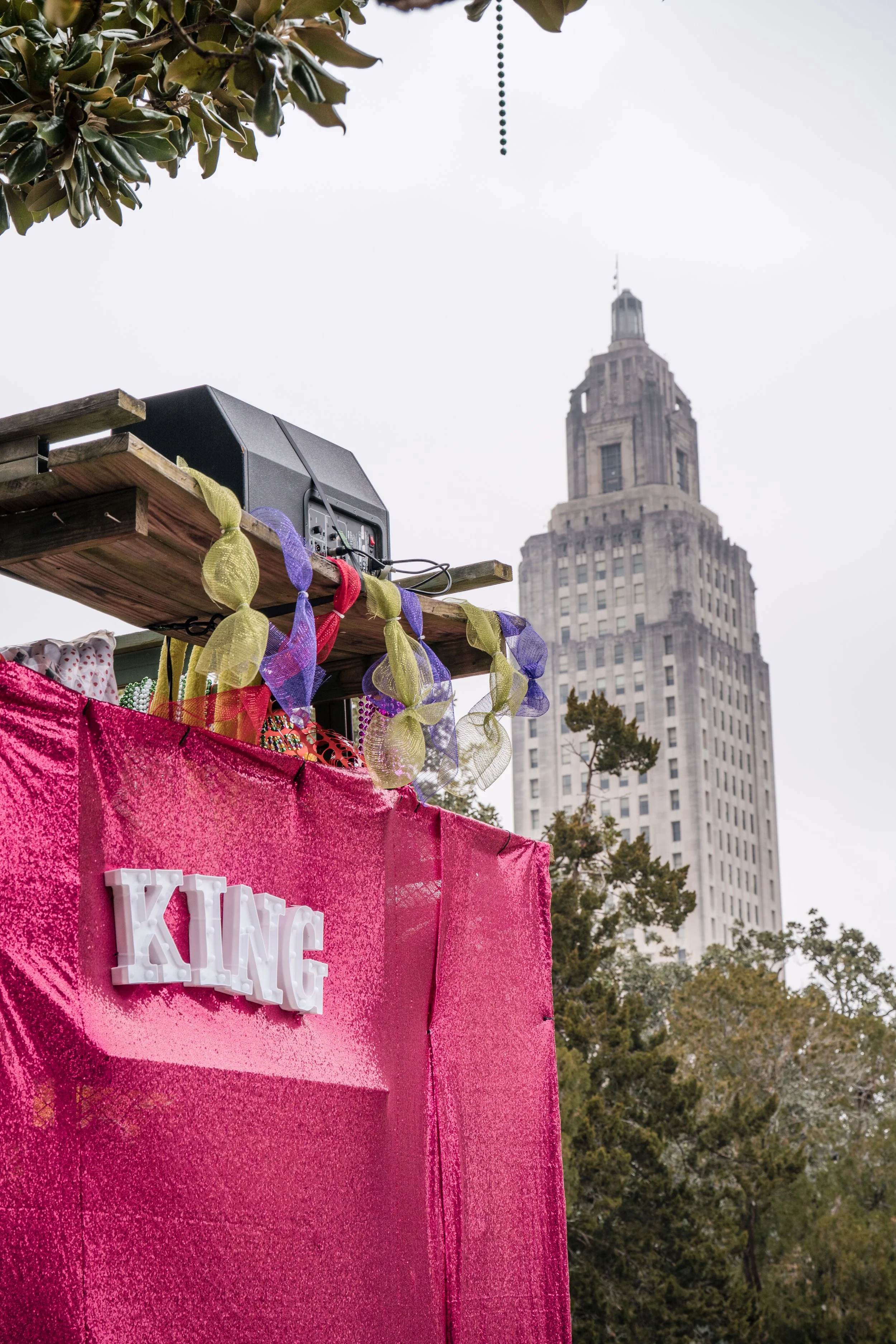 Pink glittery float with the word 'KING' displayed on it, set against a cityscape with a tall art deco style building in the background. There are decorative ribbons and a speaker on top of the float.