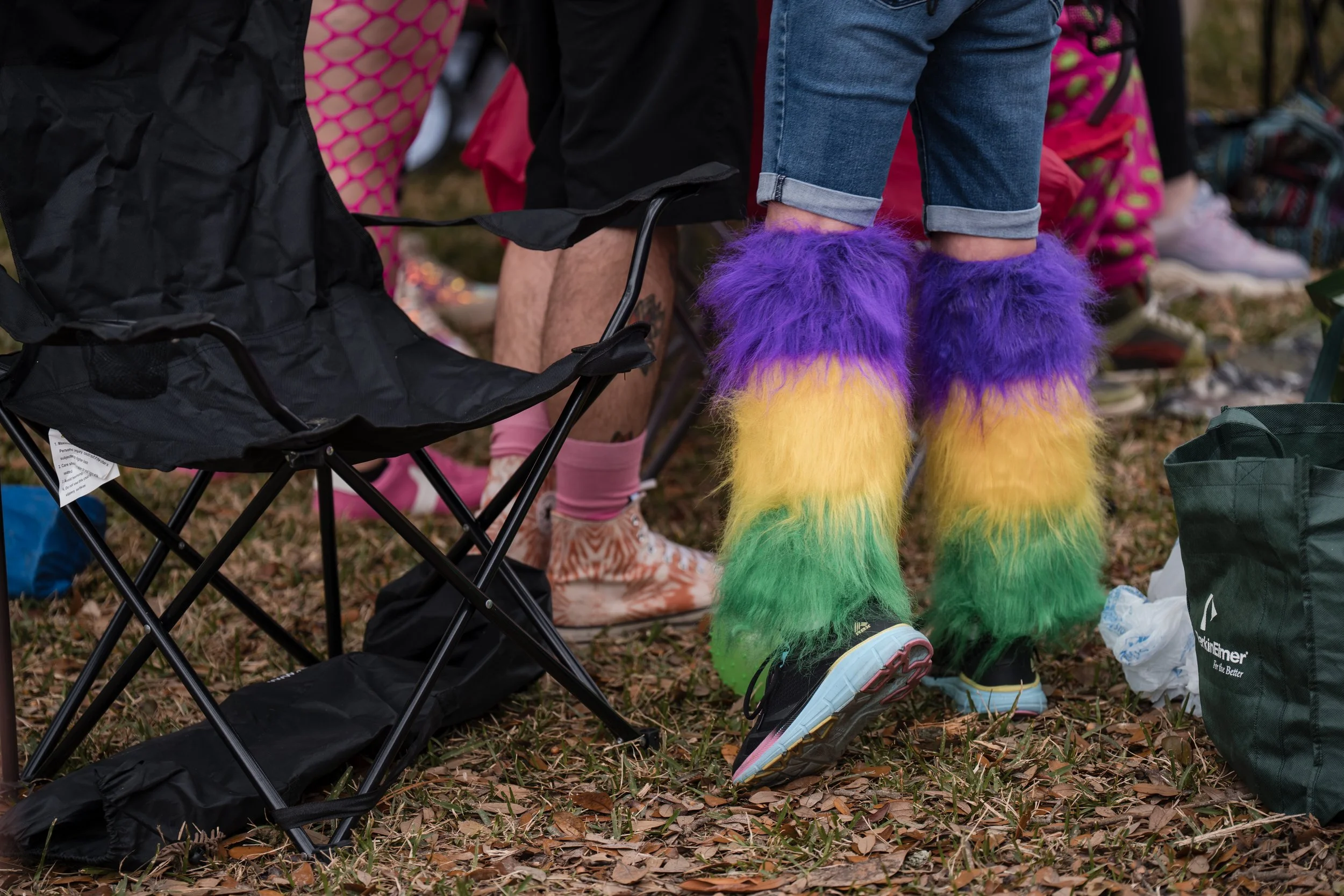 Person wearing rainbow colored furry leg warmers and sneakers, sitting on a foldable chair outdoors, with other people in the background and camping gear around.