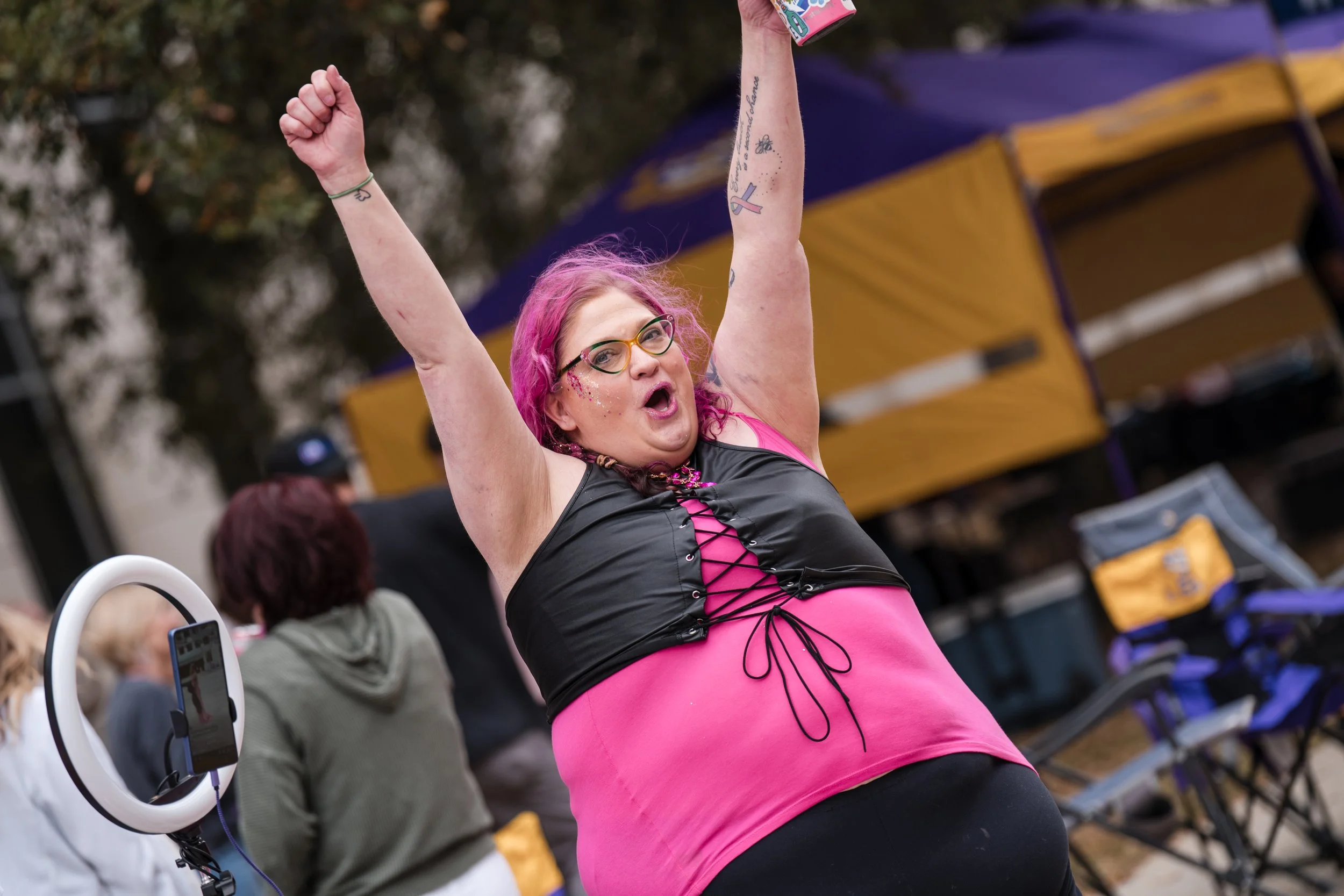 A woman with pink hair and glasses celebrates with arms raised at an outdoor event, wearing a pink and black outfit.