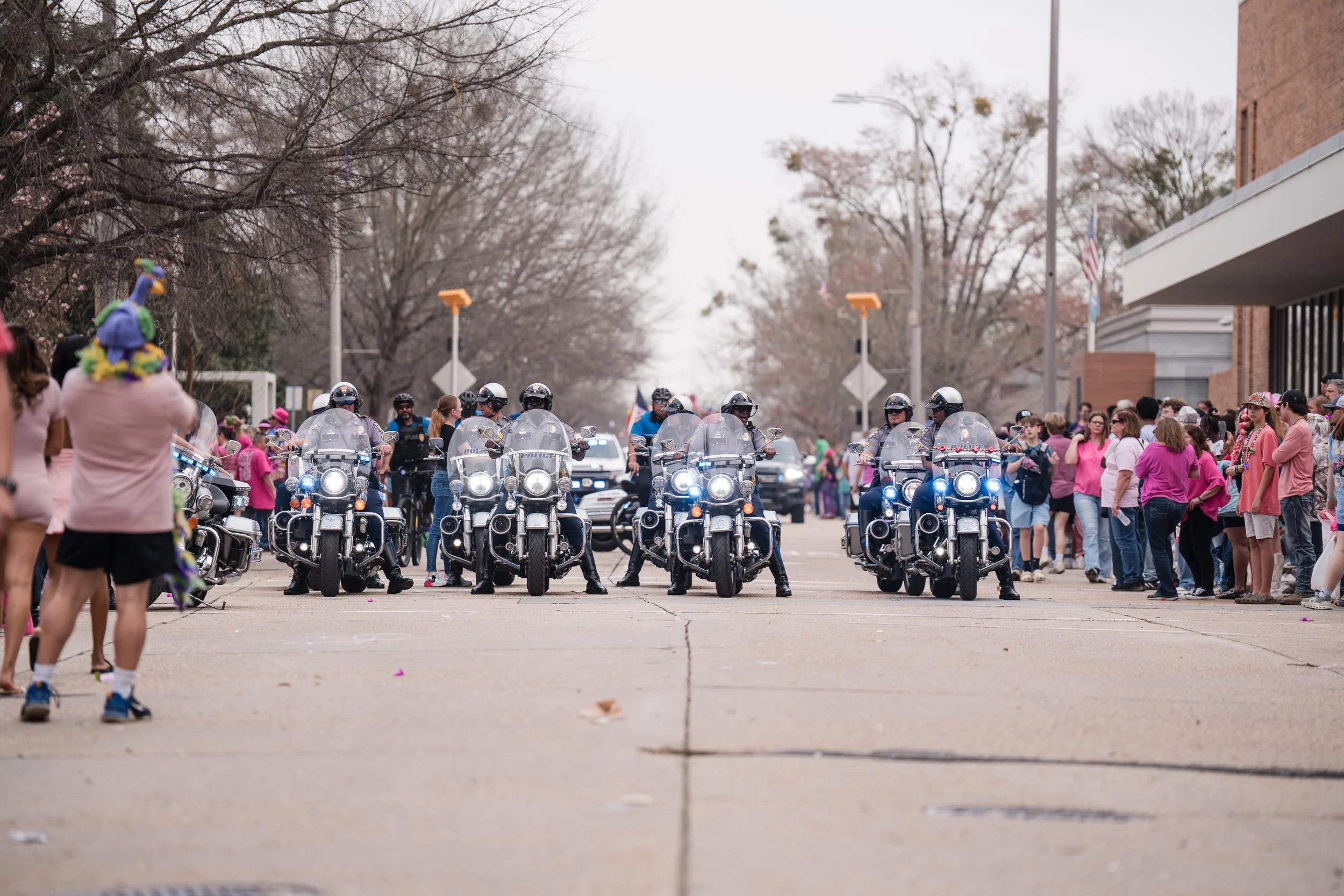 Police officers on patrol motorcycles in a parade, with a crowd of spectators lining the street on either side, some dressed in pink and various colors, during a community event or celebration.