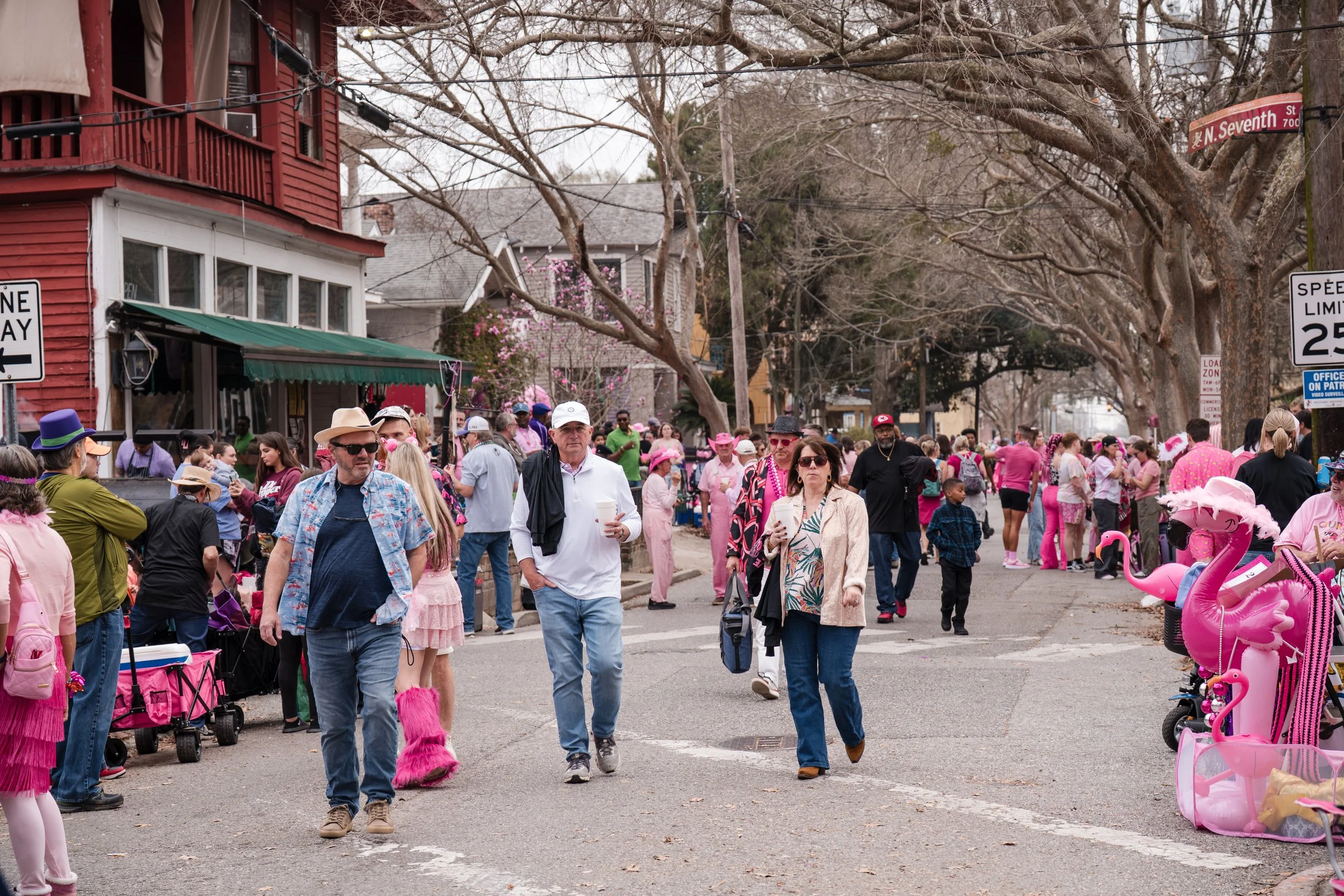 People walking on a city street decorated for a pink-themed event, with some wearing pink clothing and accessories, pink balloons and inflatable flamingos on the sidewalk, and leafless trees lining the street.