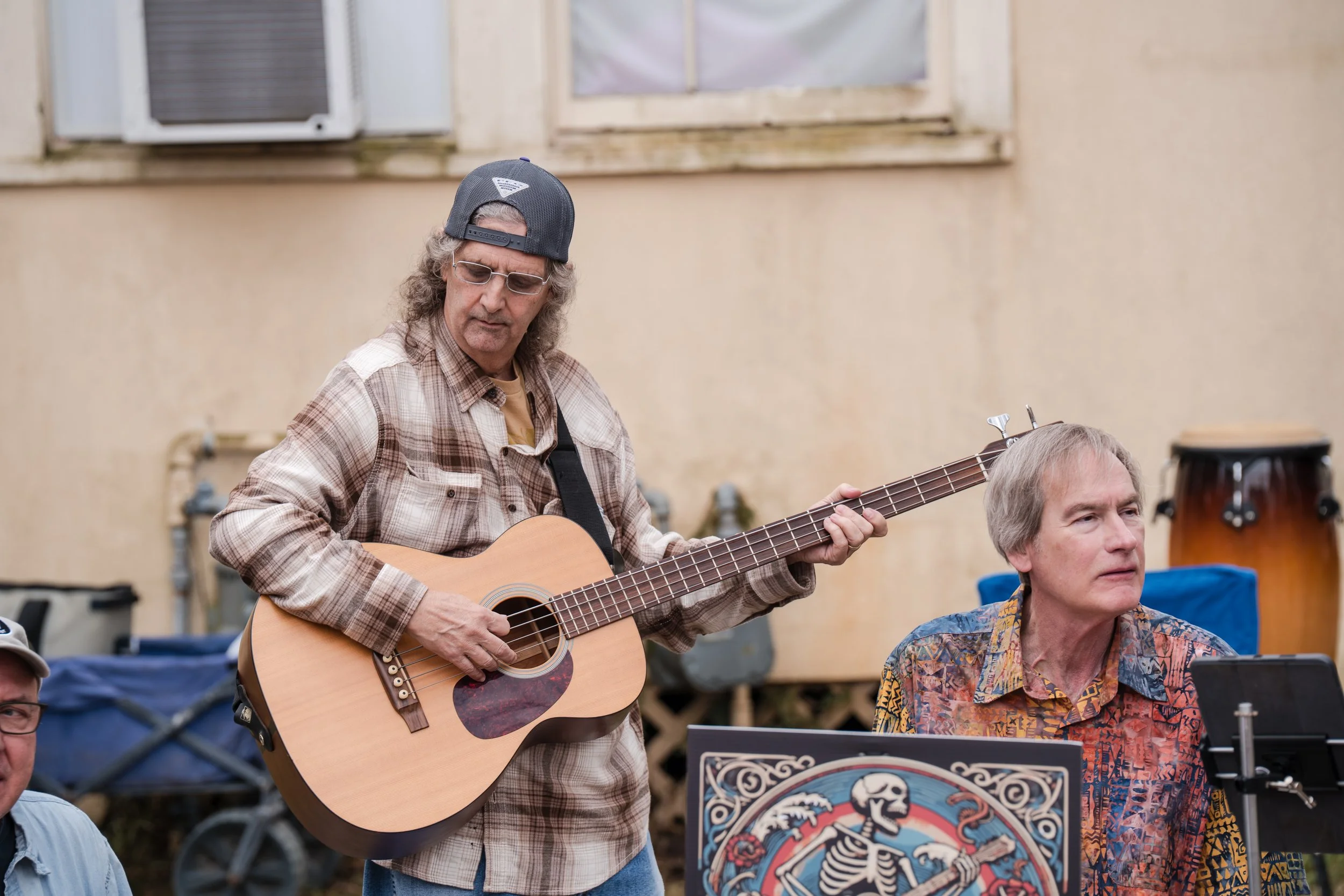 Two men perform outdoors, one playing an acoustic guitar and the other sitting nearby, with a colorful skeleton artwork in front of them.