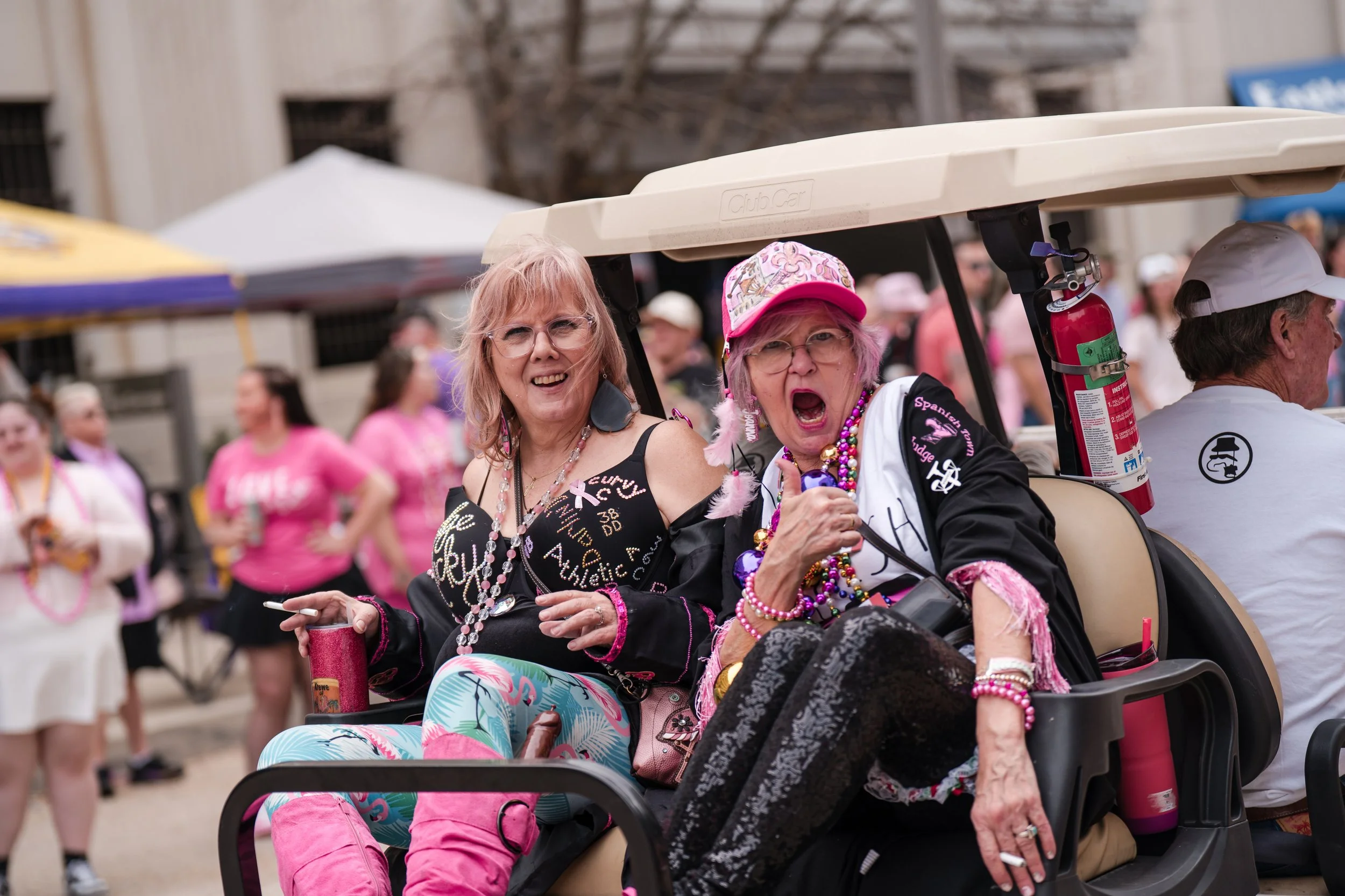 Two women, dressed in colorful and festive clothing, sitting on a golf cart at a pride event. One woman is smiling while the other is shouting or singing, surrounded by people in pink outfits and rainbow-themed accessories.