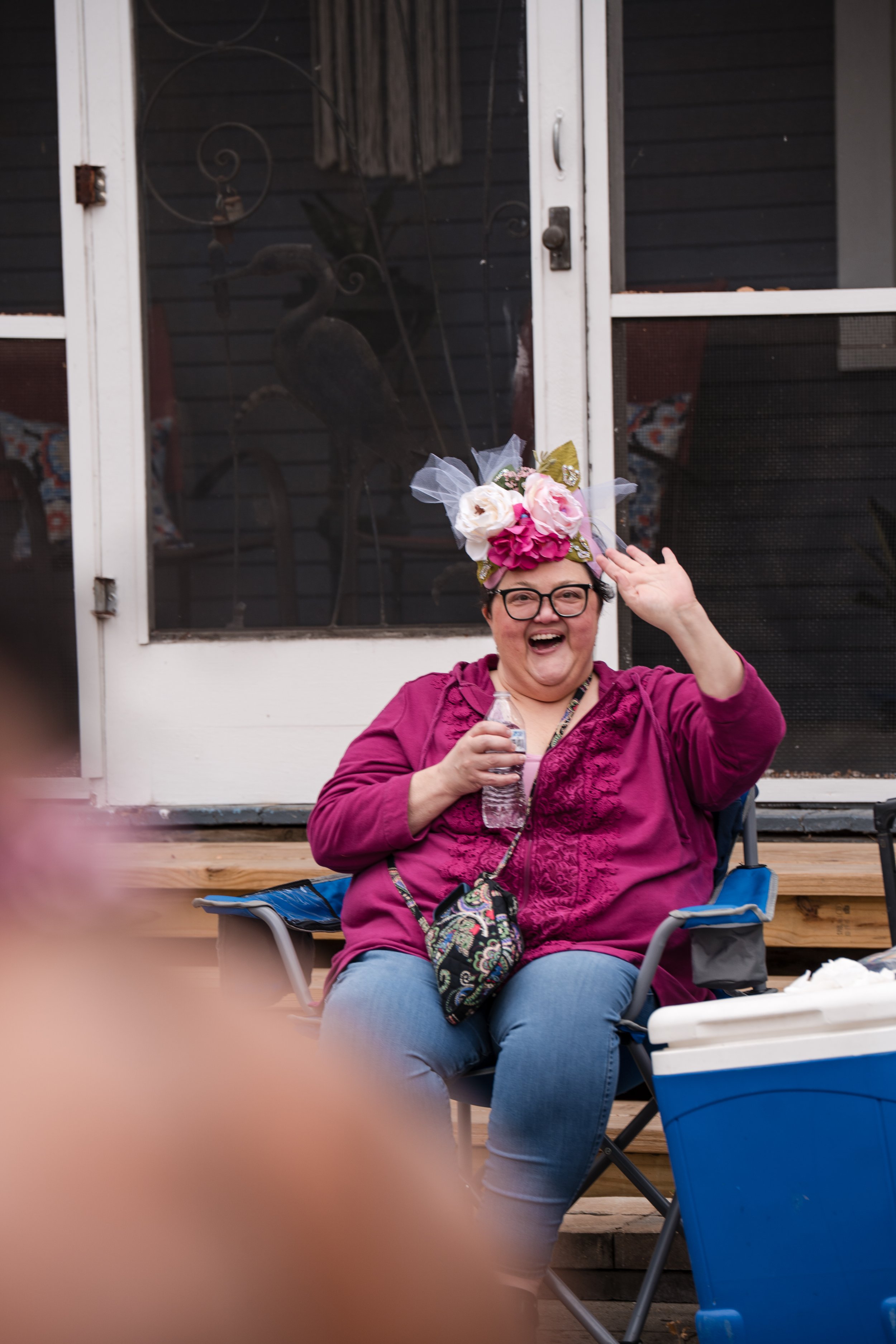 A cheerful woman in glasses and a purple hoodie sitting outside, wearing a floral hat, holding a water bottle, waving, and smiling.