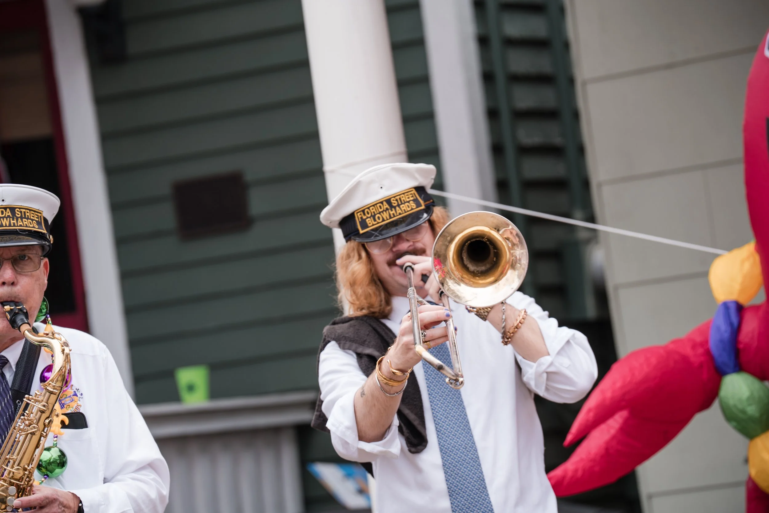 A woman playing a trumpet during an outdoor parade, wearing a white shirt, white cap, and sunglasses, with a colorful float or decoration visible on the right side.
