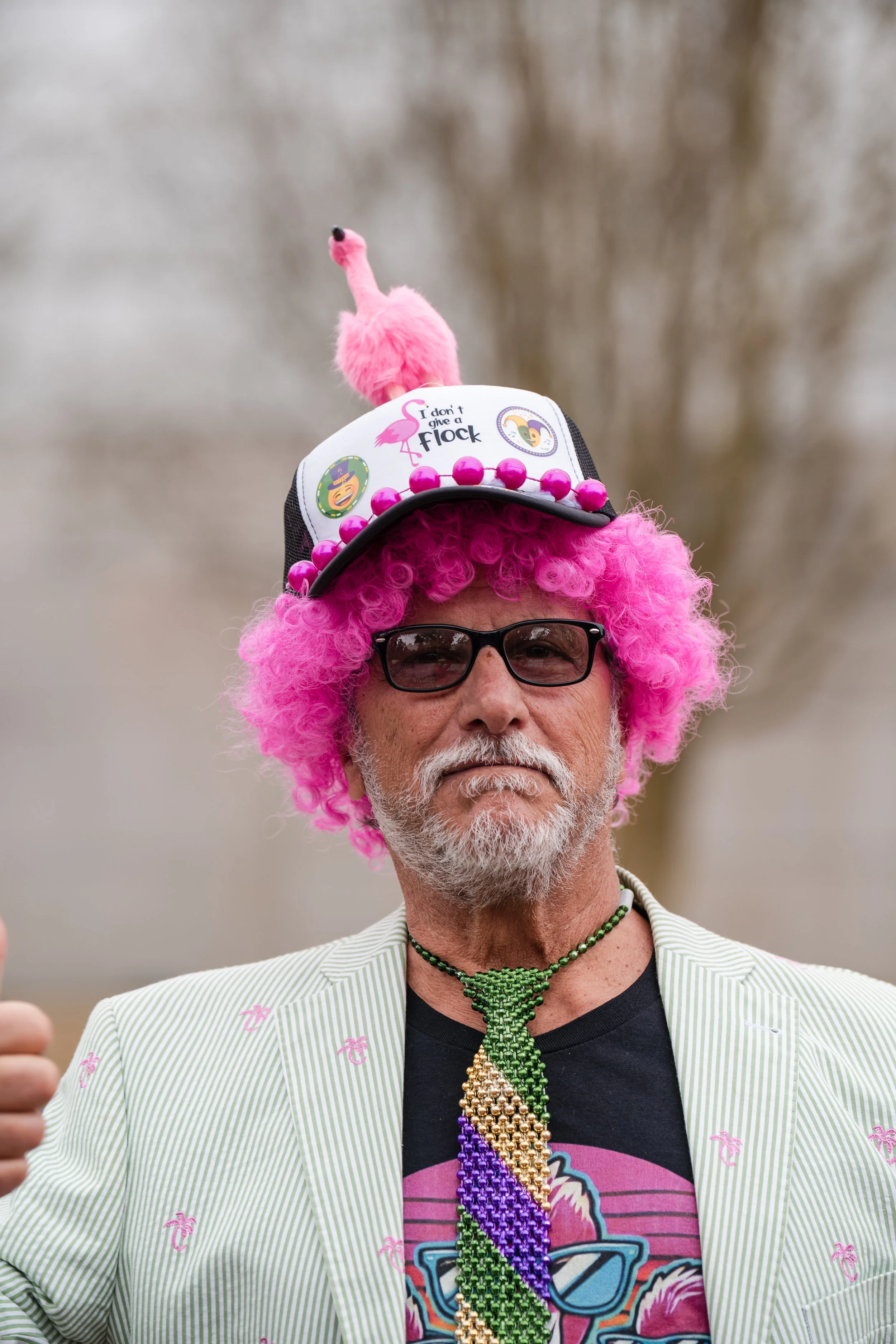 Man with a pink curly wig, wearing sunglasses, a light green striped blazer with pink palm tree prints, and a beaded rainbow necklace, giving a thumbs up. He is also wearing a hat decorated with pink beads, a plush flamingo, and patches that says 'I 
